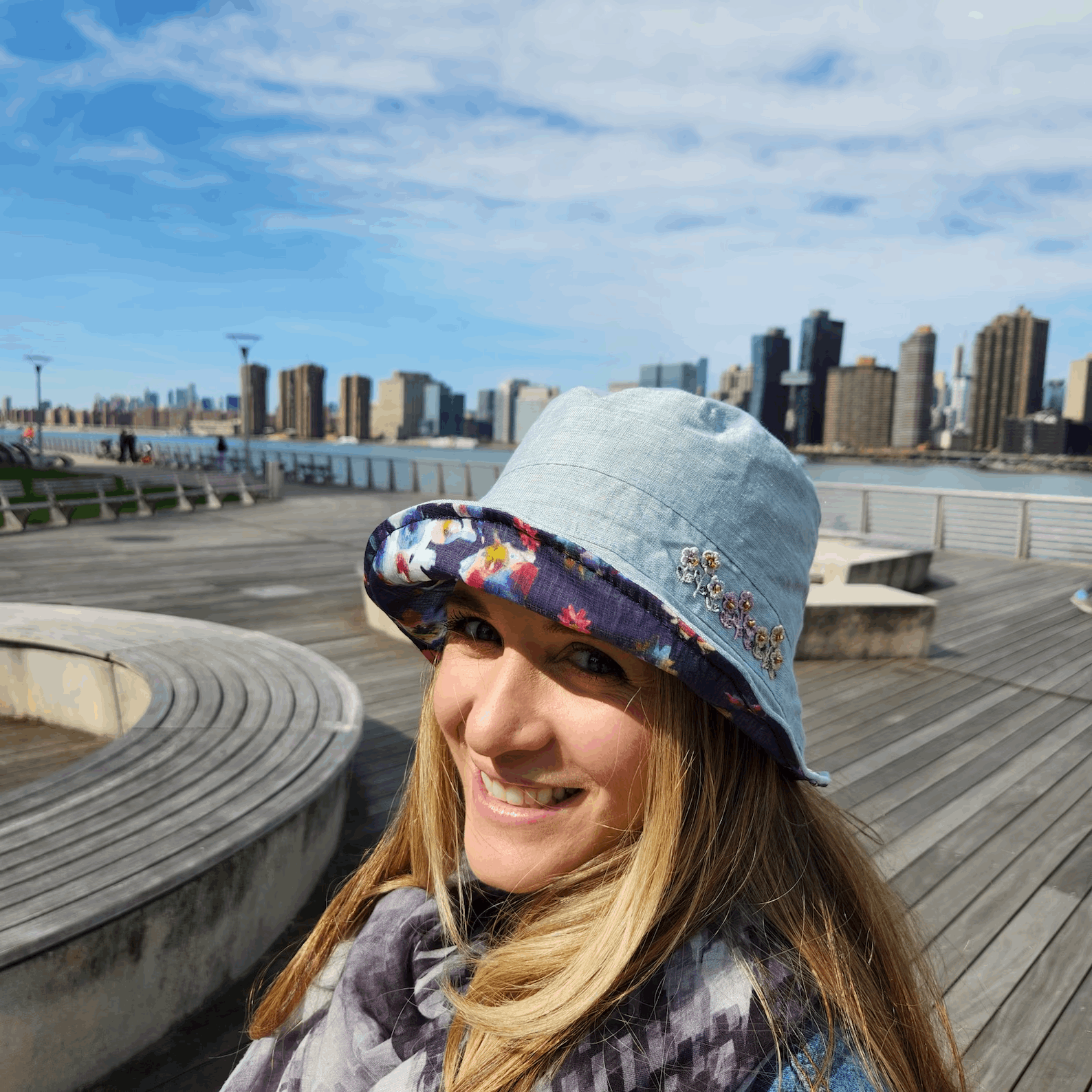 Woman in a light blue floral linen bucket-folderable hat with decorative details, smiling with a city skyline view