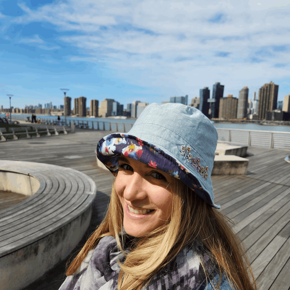 Woman in a light blue floral linen bucket-folderable hat with decorative details, smiling with a city skyline view