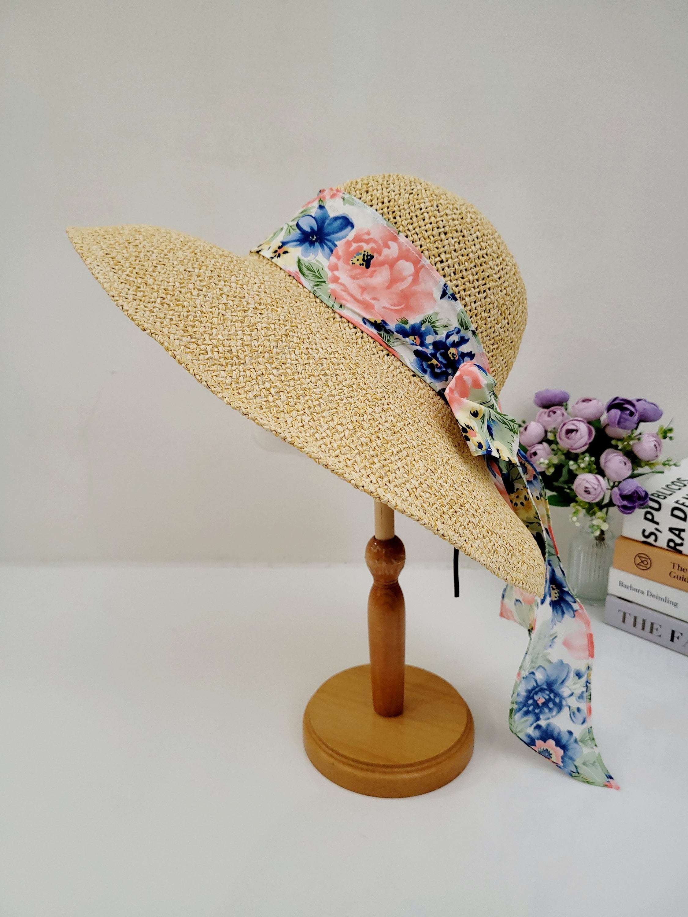 East Hampton straw sun hat with wide brim and a vibrant floral scarf accent, displayed on a wooden stand.