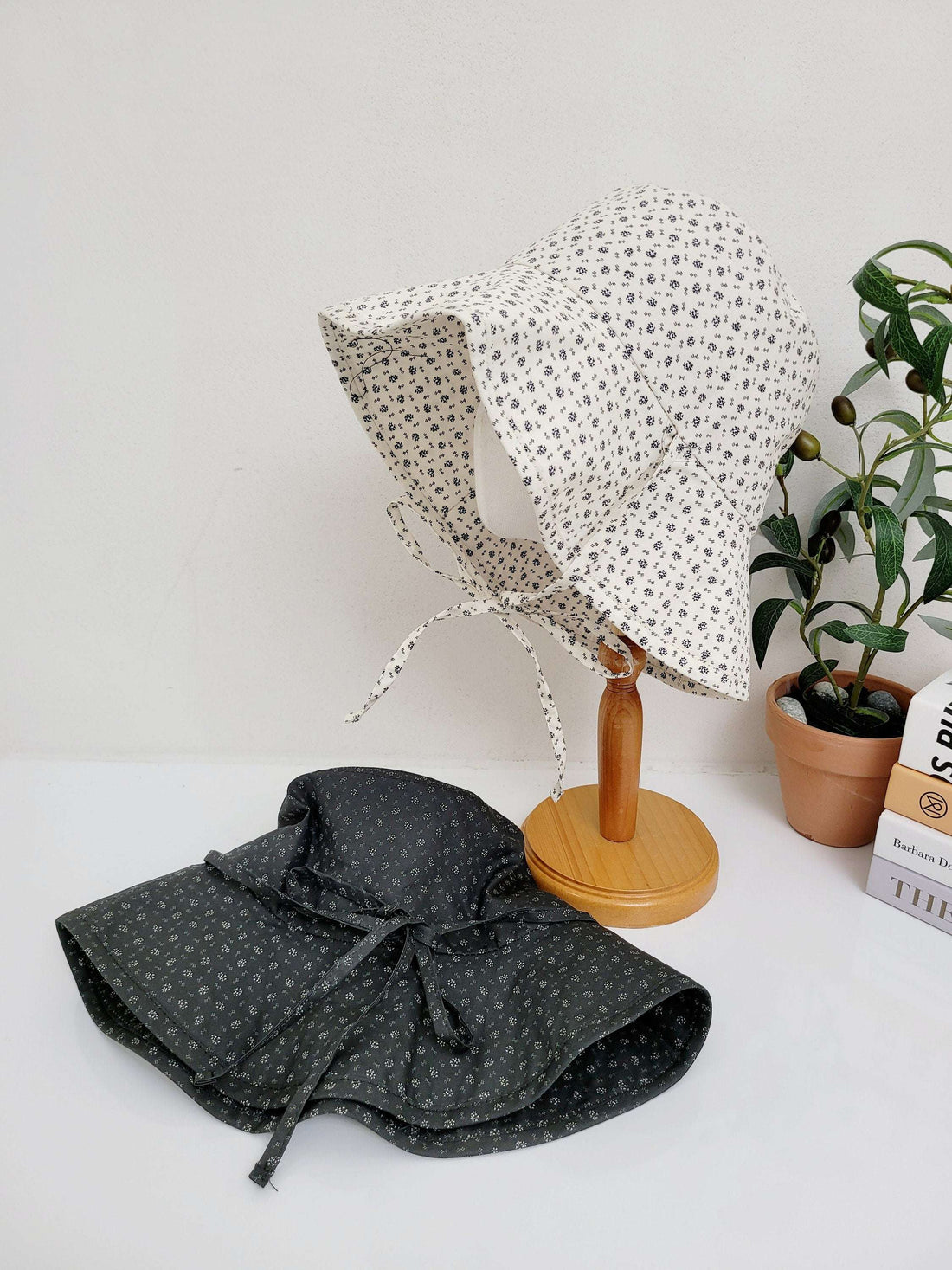 Two light weight wide brim hat sunbonnets: one cream with black floral print on a stand, and a black with white floral print lying flat with tie closure.