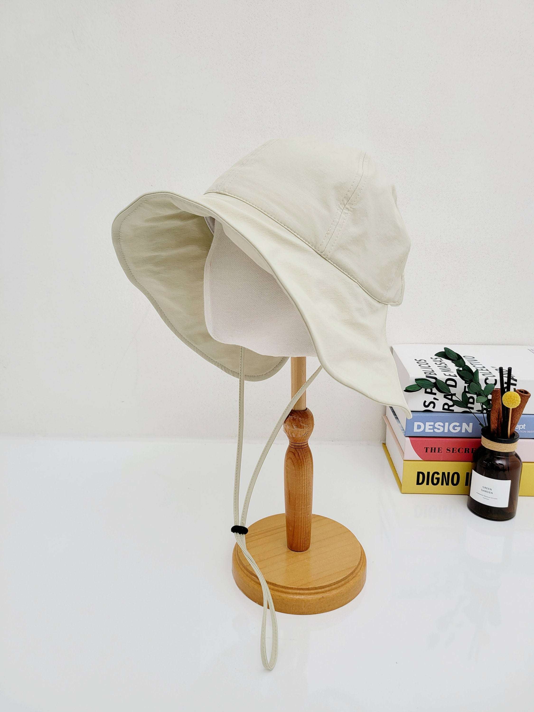 SUMMER LARGE BRIM HAT, a beige sun hat with a wide brim, displayed on a wooden stand. Features a chin strap for secure wear
