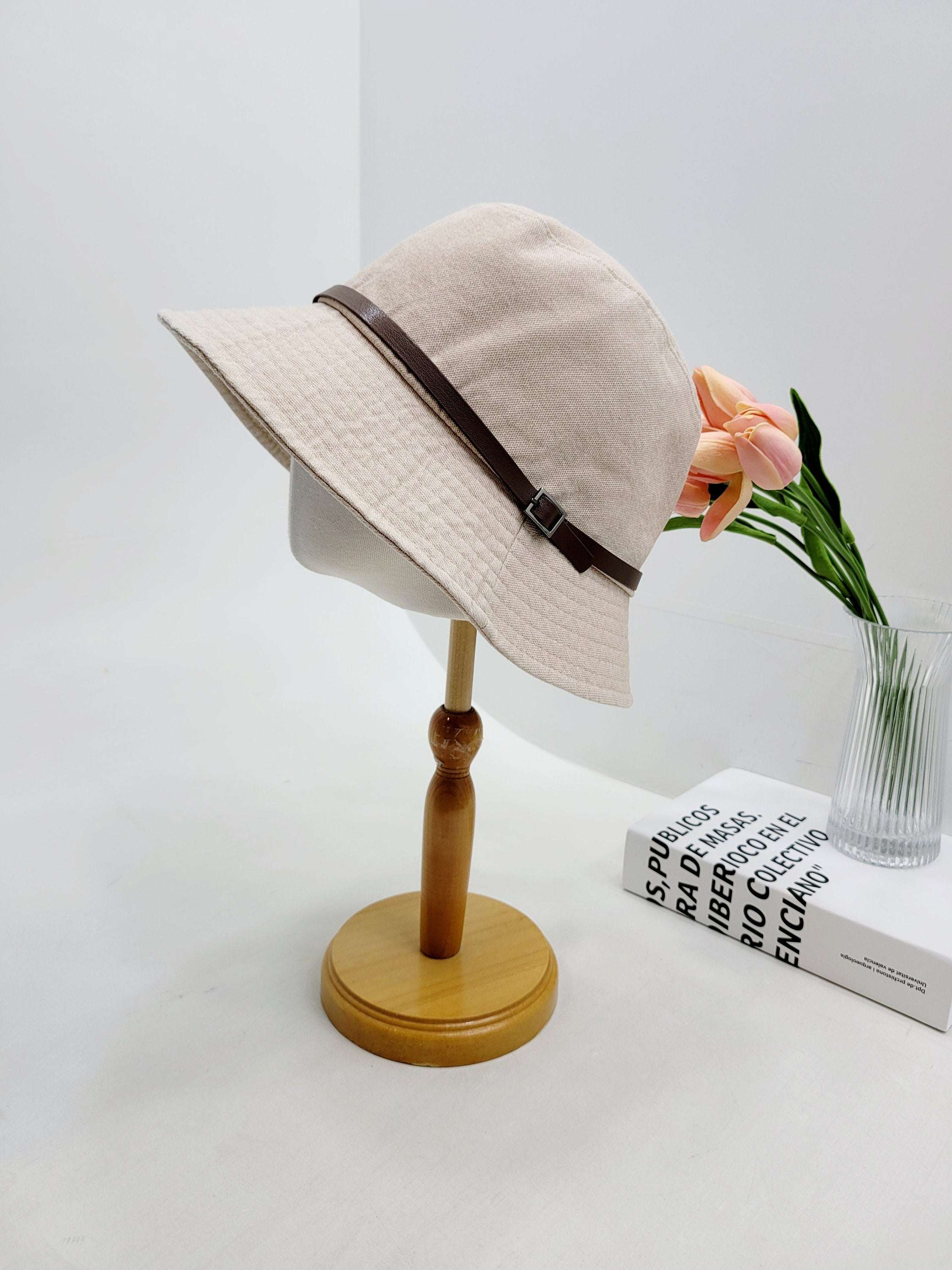 Beige bucket hat, the miles, with a brown leather strap and buckle detail, displayed on a wooden stand.