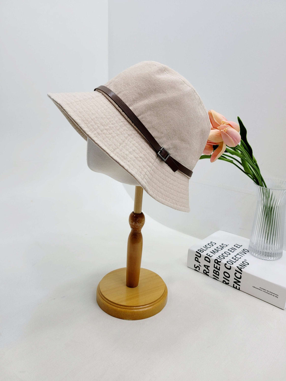 Beige bucket hat, the miles, with a brown leather strap and buckle detail, displayed on a wooden stand.