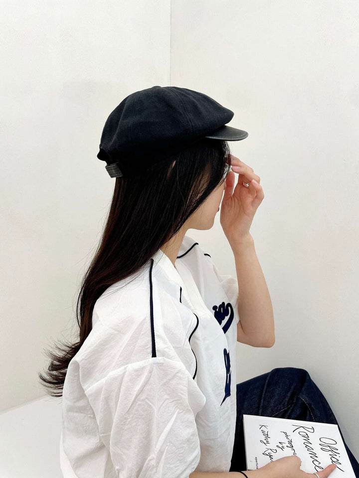 Woman in profile wearing a black lana beret and white shirt, holding a book. The beret has a classic, slightly puffed design.