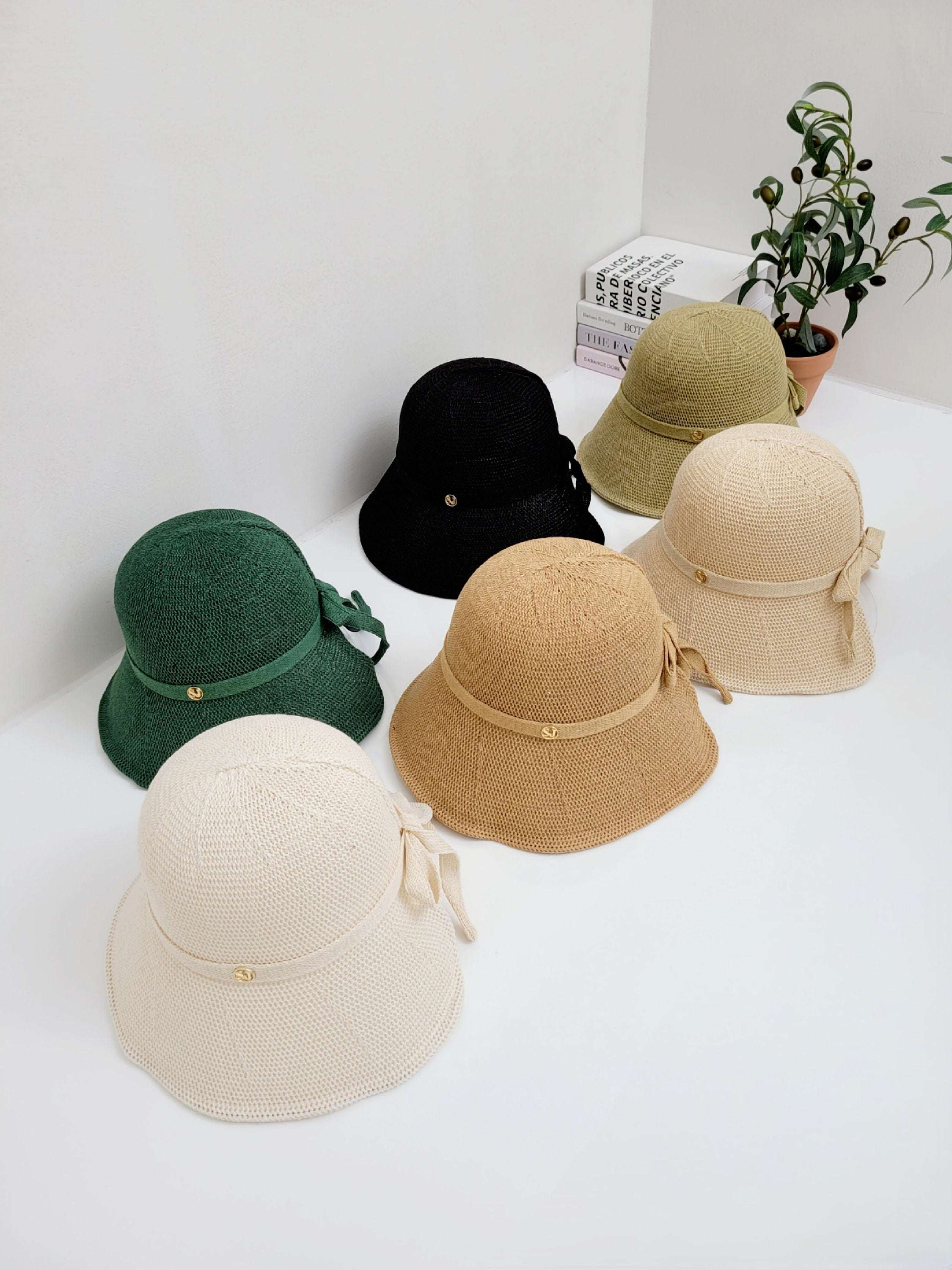 Six knitted sun hats in cream, tan, green, olive, black, and beige, each with a ribbon detail, arranged on a white surface