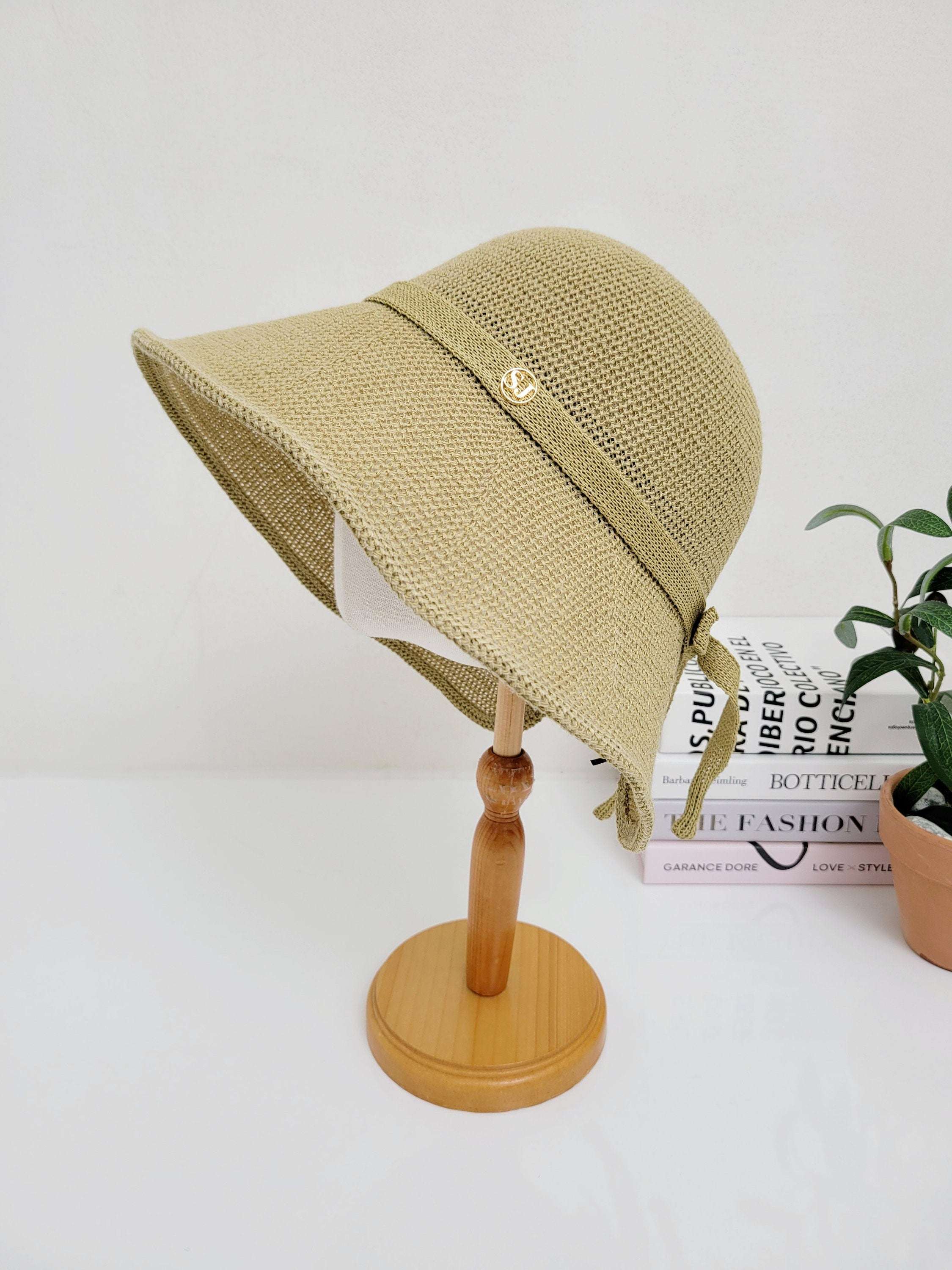 Tan woven sun hat SJ with ribbon tie and smiley detail, on a wooden stand. Books and plant in background