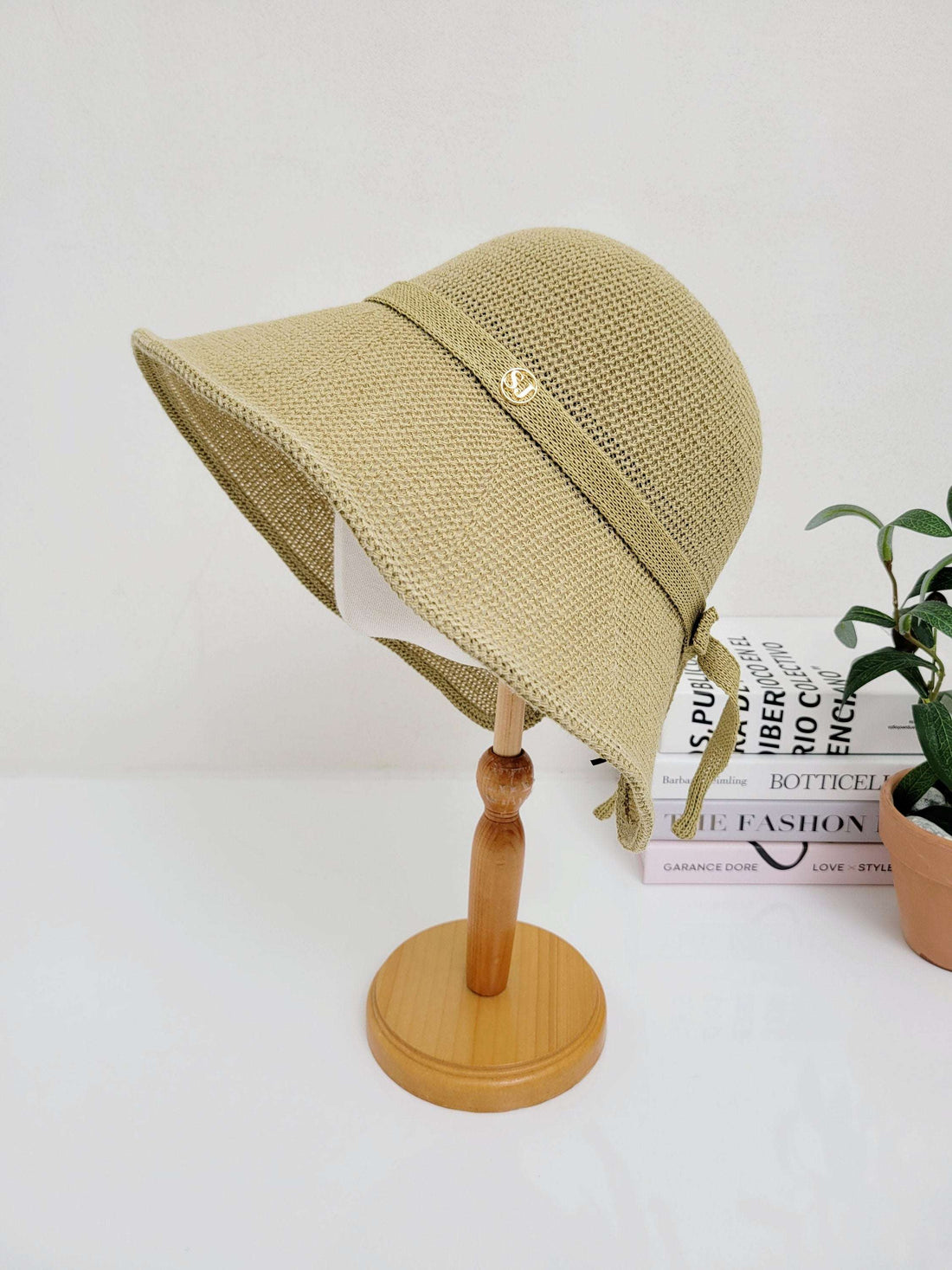 Tan woven sun hat SJ with ribbon tie and smiley detail, on a wooden stand. Books and plant in background