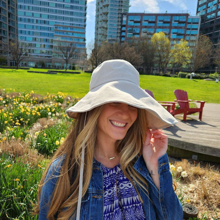 Woman in a cotton linen wide brim-solid hat, smiling in a sunny park with flowers. Light beige sun hat with denim jacket.