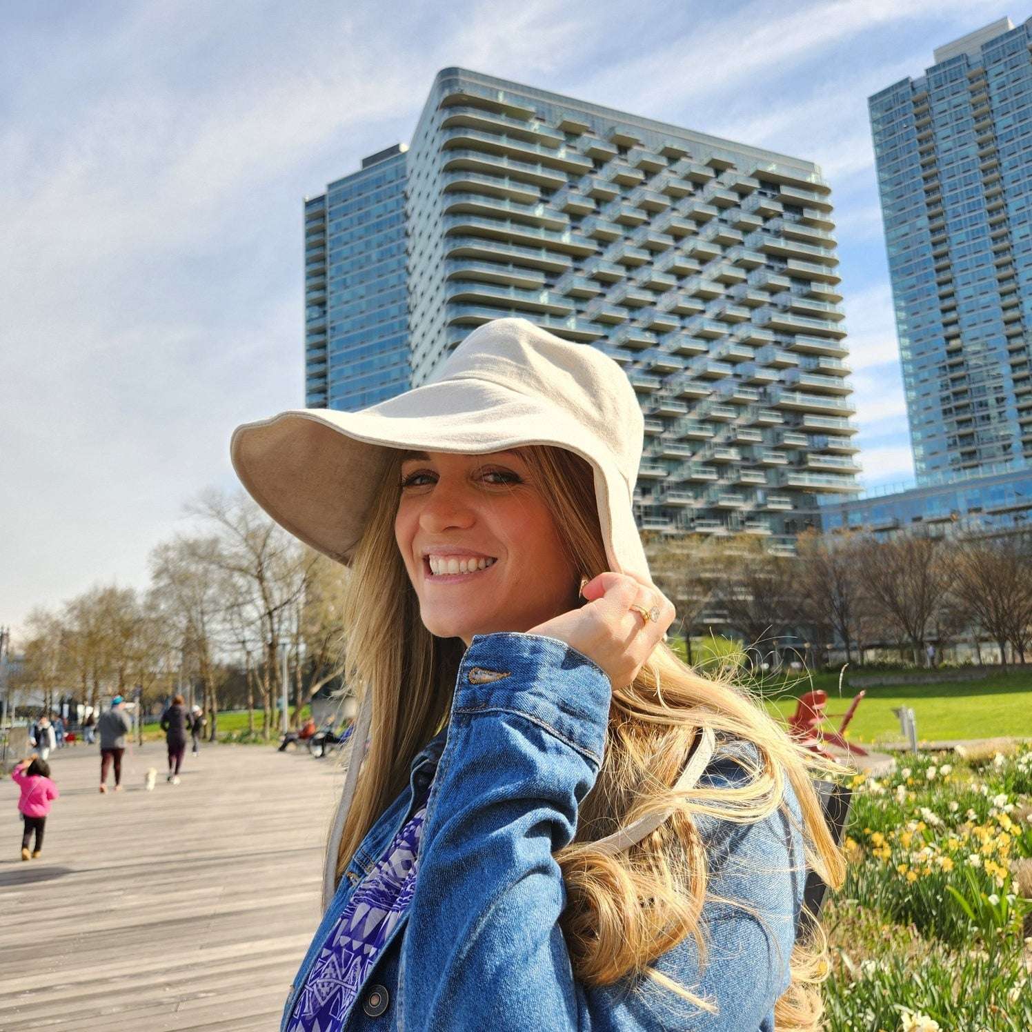 Woman in cotton linen wide brim-solid hat, denim jacket, and patterned top smiles outdoors. The wide brim provides sun protection.