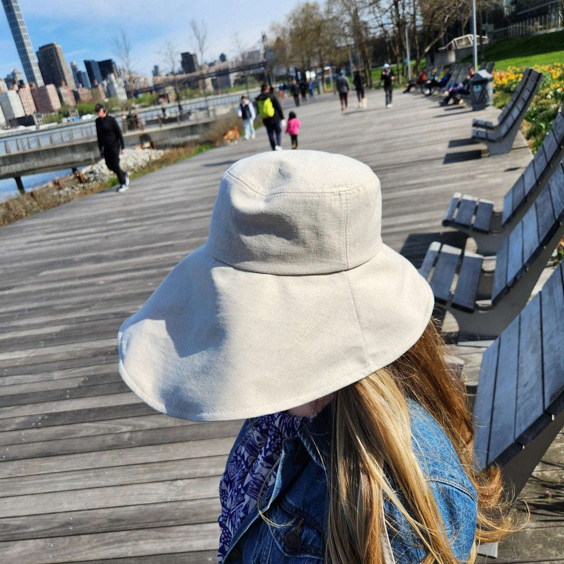Woman wears a cotton linen wide brim-solid hat in off-white, sitting on a boardwalk with city views.
