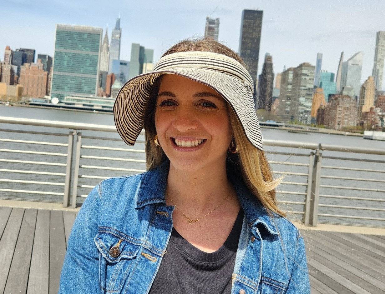 Woman smiling, wearing a lic suncap stripe visor with a black and white pattern, denim jacket, and gold jewelry, city skyline background