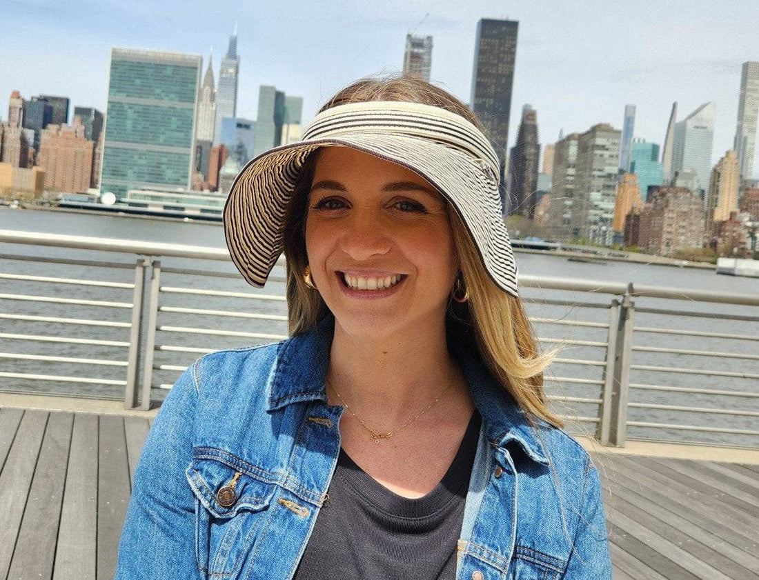 Woman smiling, wearing a lic suncap stripe visor with a black and white pattern, denim jacket, and gold jewelry, city skyline background
