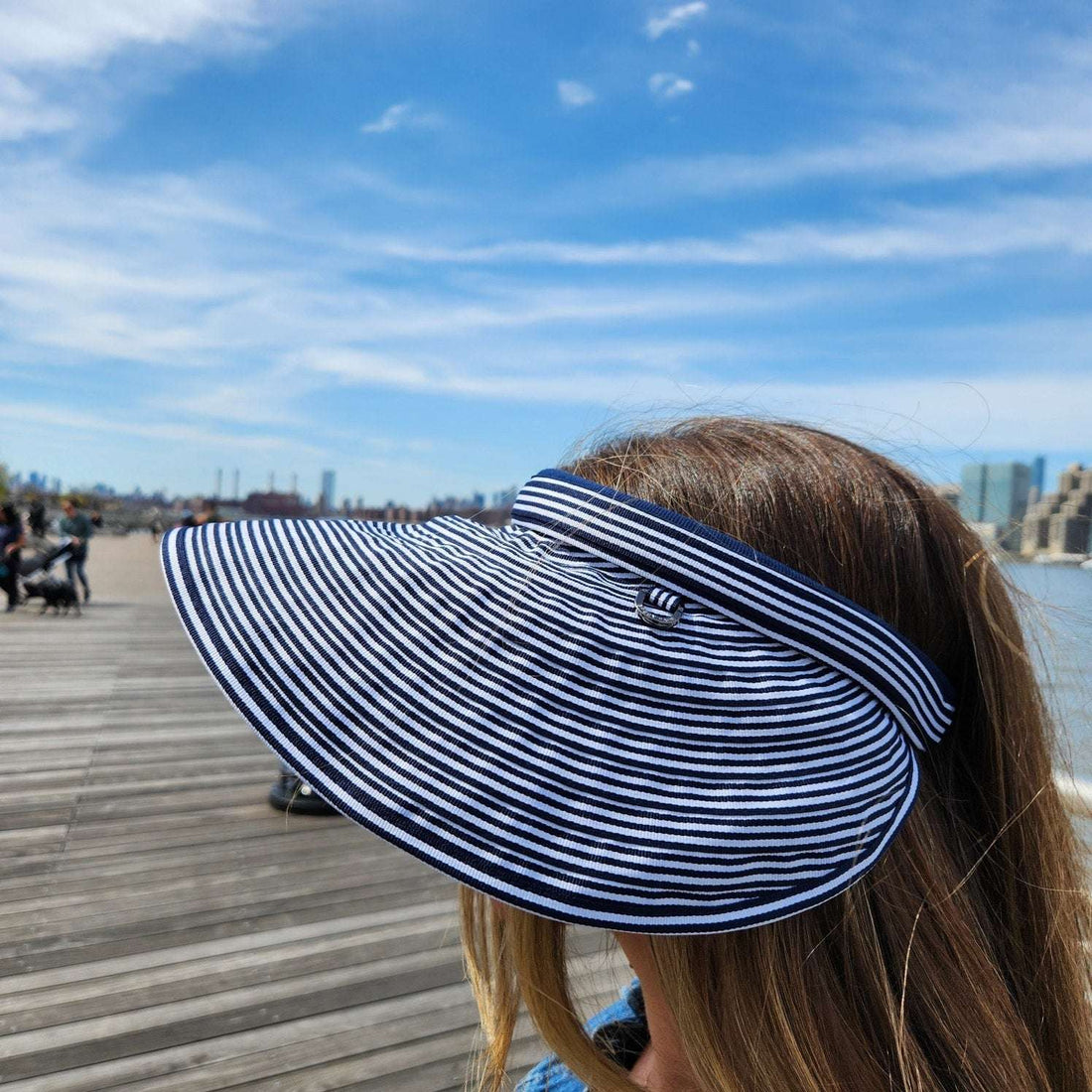 Woman wearing a lic suncap stripe visor with blue and white stripes, standing on a wooden boardwalk with a city skyline in the background.