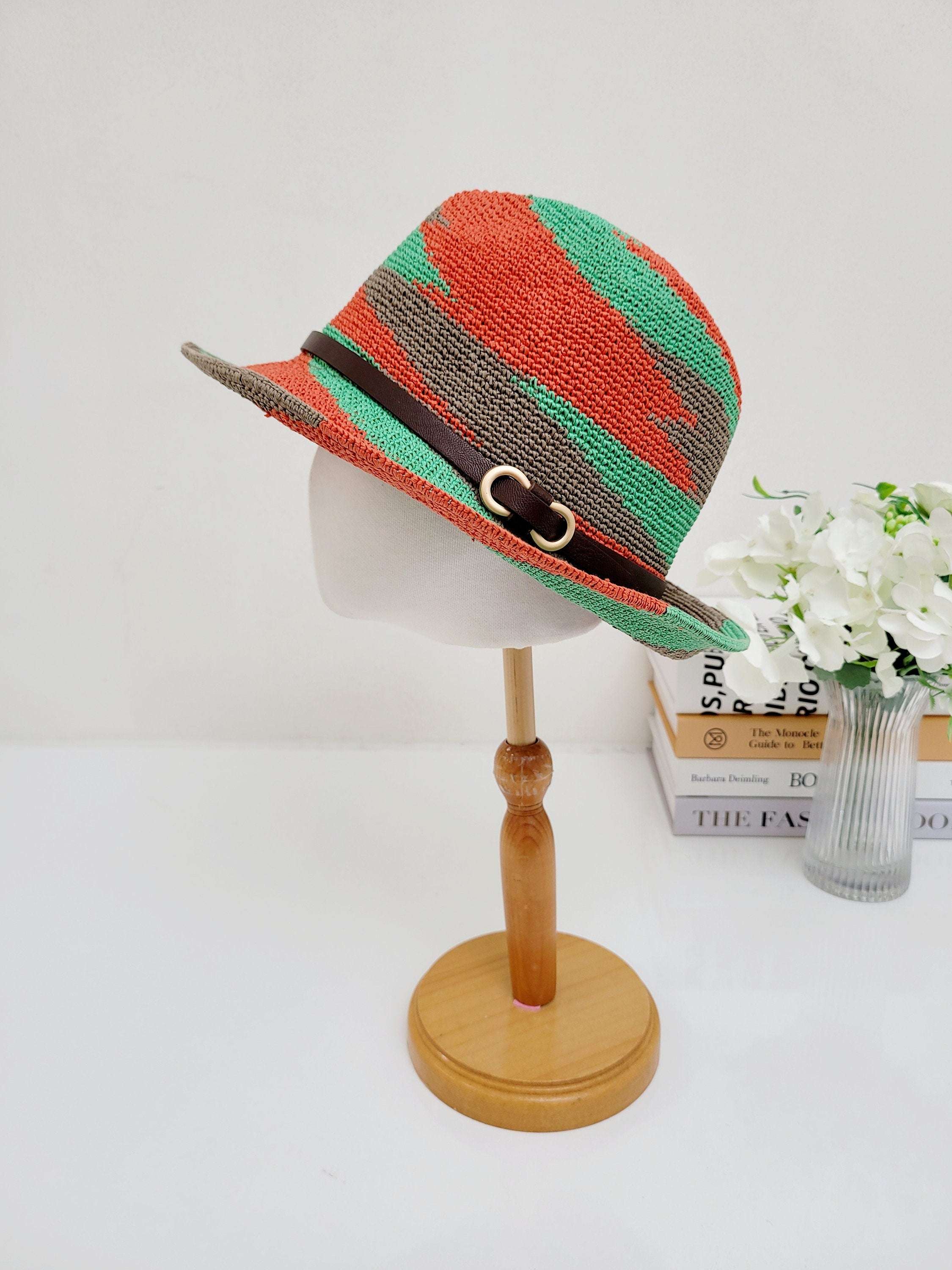 serengeti hat: woven fedora in red, green, and brown with a brown faux leather band and gold buckle, displayed on a wooden stand.