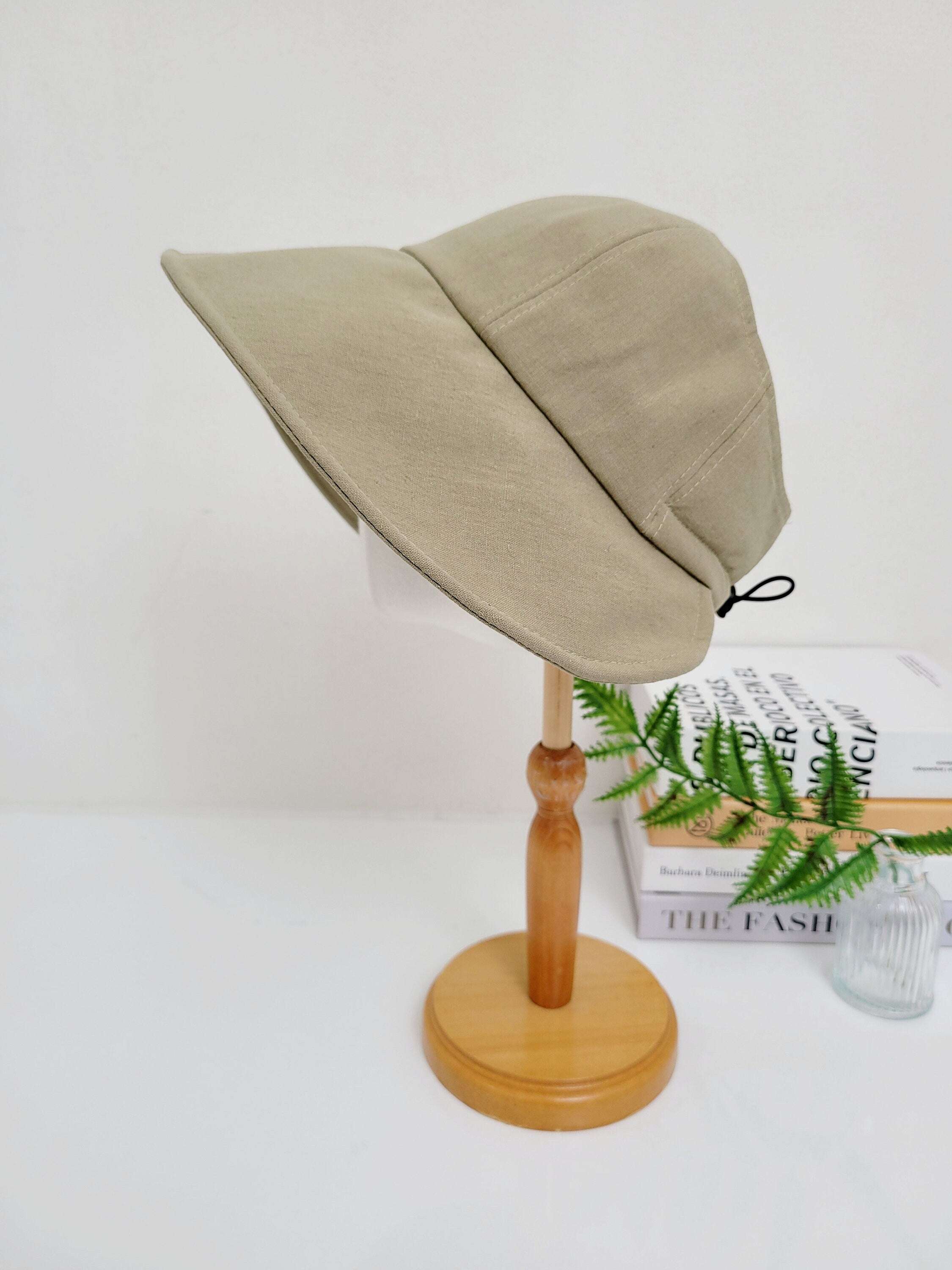 Beige linen sun hat with a wide, downturned brim, displayed on a wooden stand. A stylish and practical summer accessory.