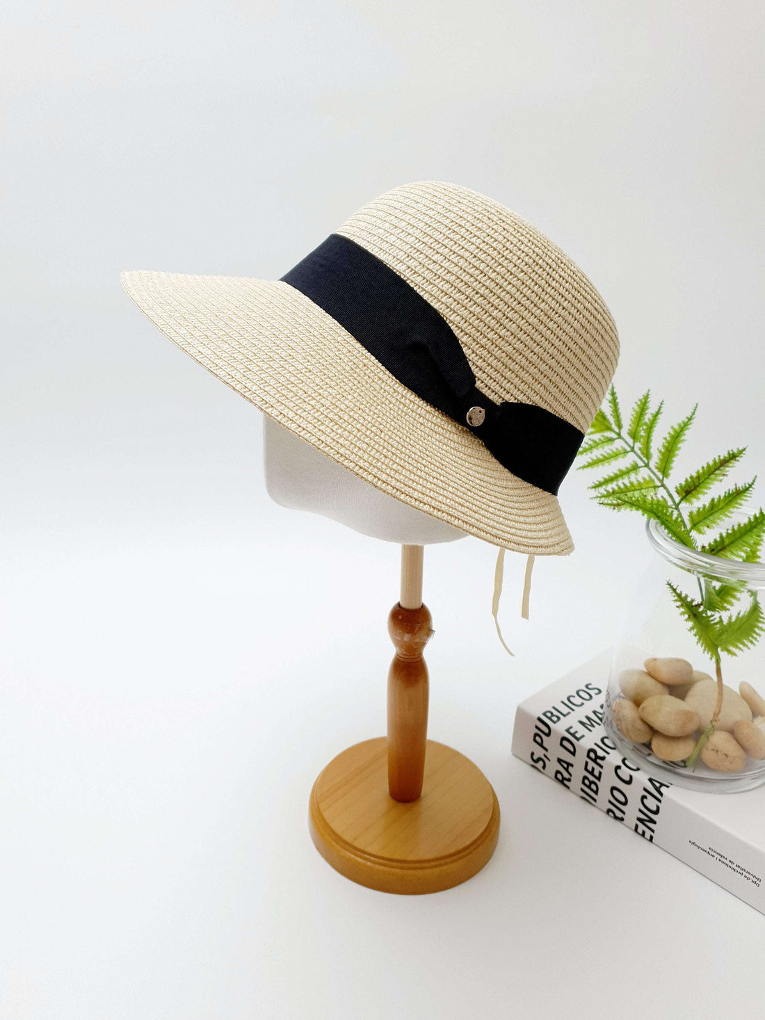 ISABELLE: Beige straw sun hat with a stylish black ribbon accent and a decorative button, displayed on a wooden stand.