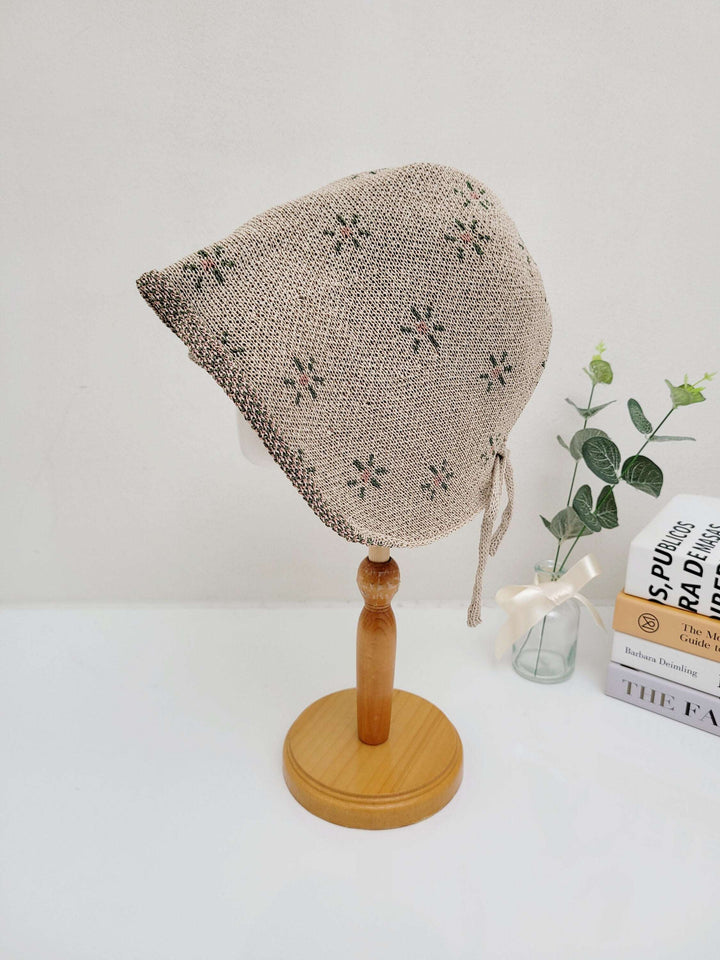 emma: Beige knitted bonnet with a delicate floral pattern, displayed on a wooden stand with tie closures.