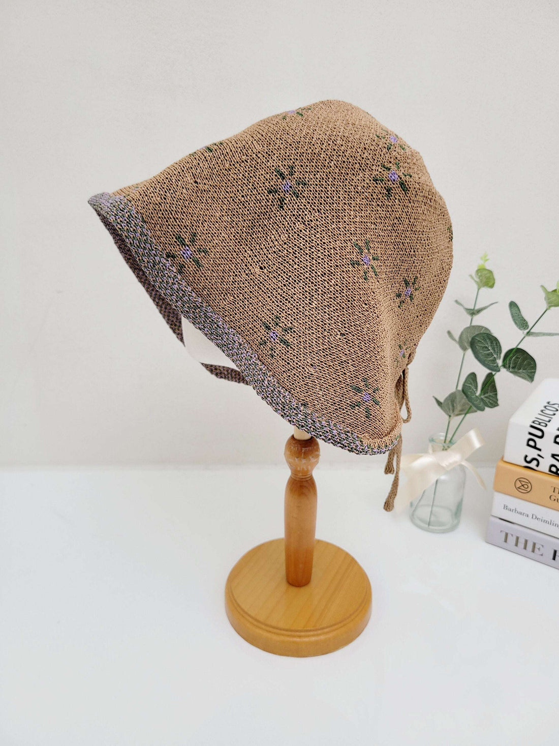 emma bonnet: Brown knit baby bonnet with a delicate floral pattern and tie closures, displayed on a wooden stand.
