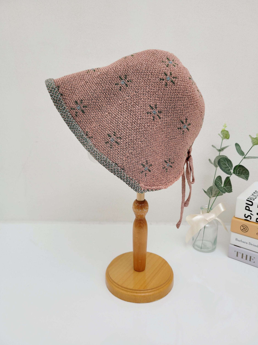 emma: Pink knit baby bonnet with delicate blue flower accents and gray trim, displayed on a wooden stand.
