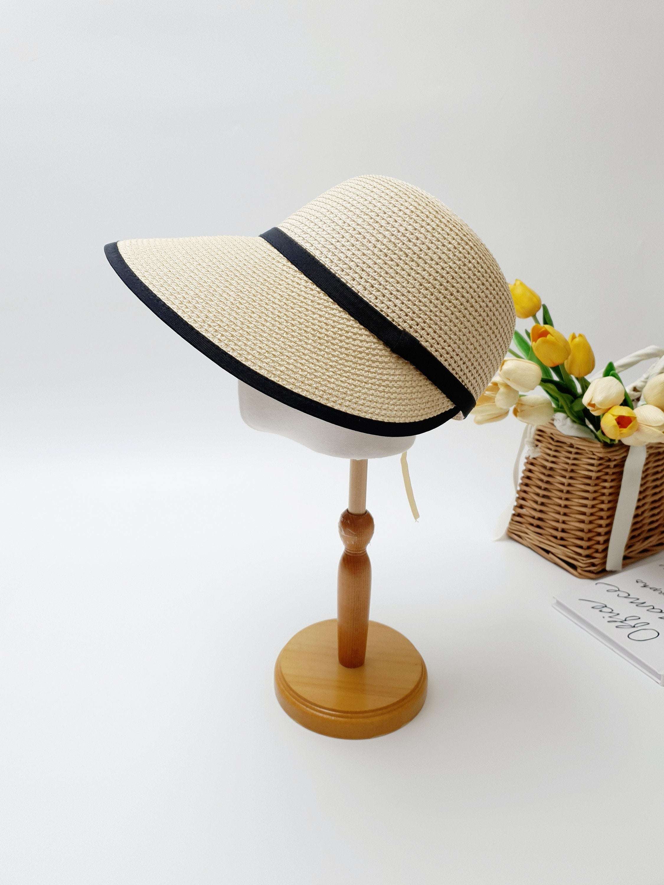Beige straw sun visor, a hepburn panama, with elegant black trim on brim and band, displayed on a wooden stand.