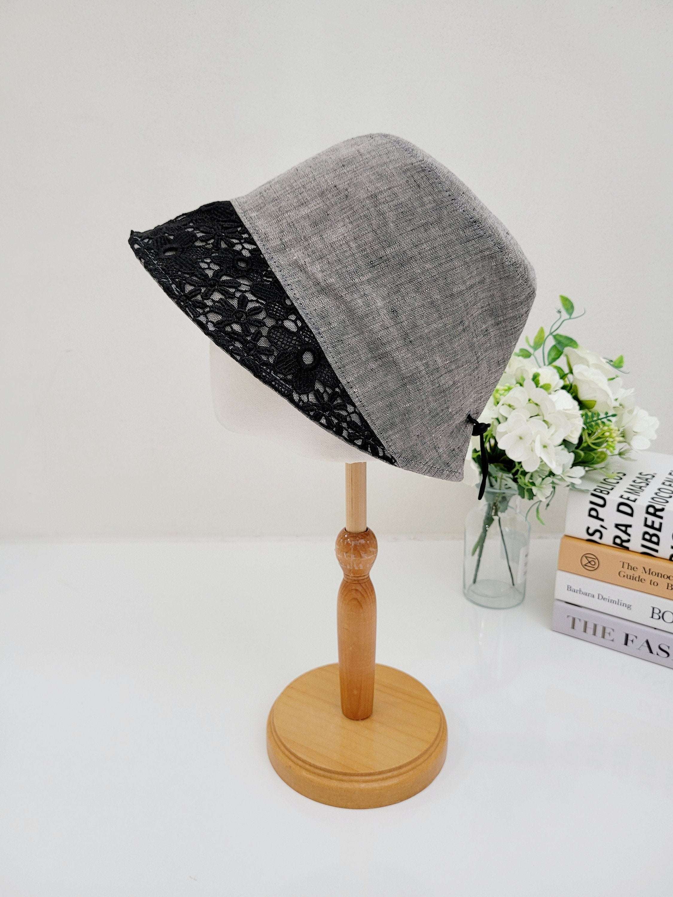 Gray linen bucket-flower eyelet hat with delicate black lace trim, displayed on a wooden stand.