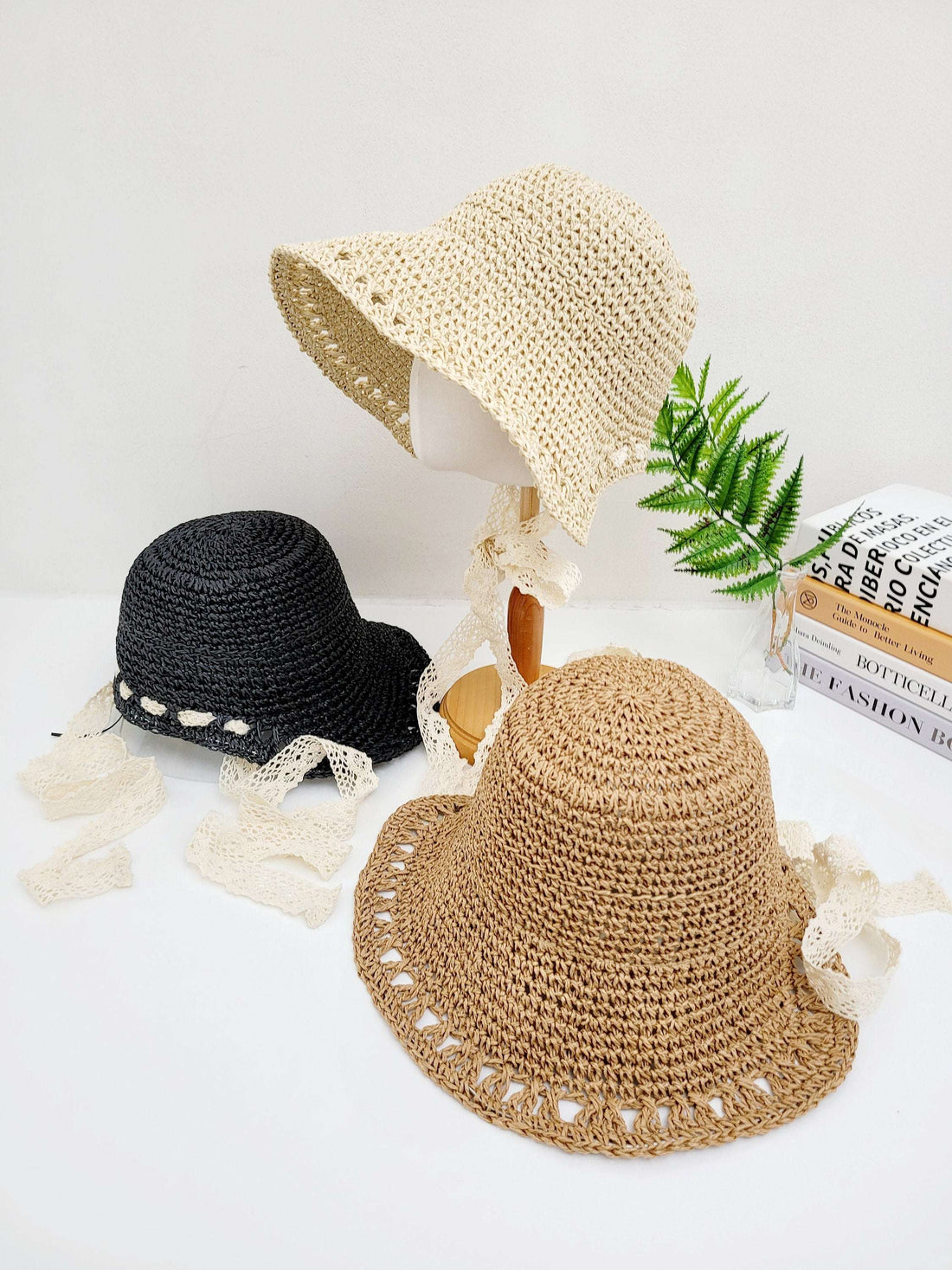 Three bora bora straw hats in beige, black, and brown with delicate lace ties, arranged on a white surface.