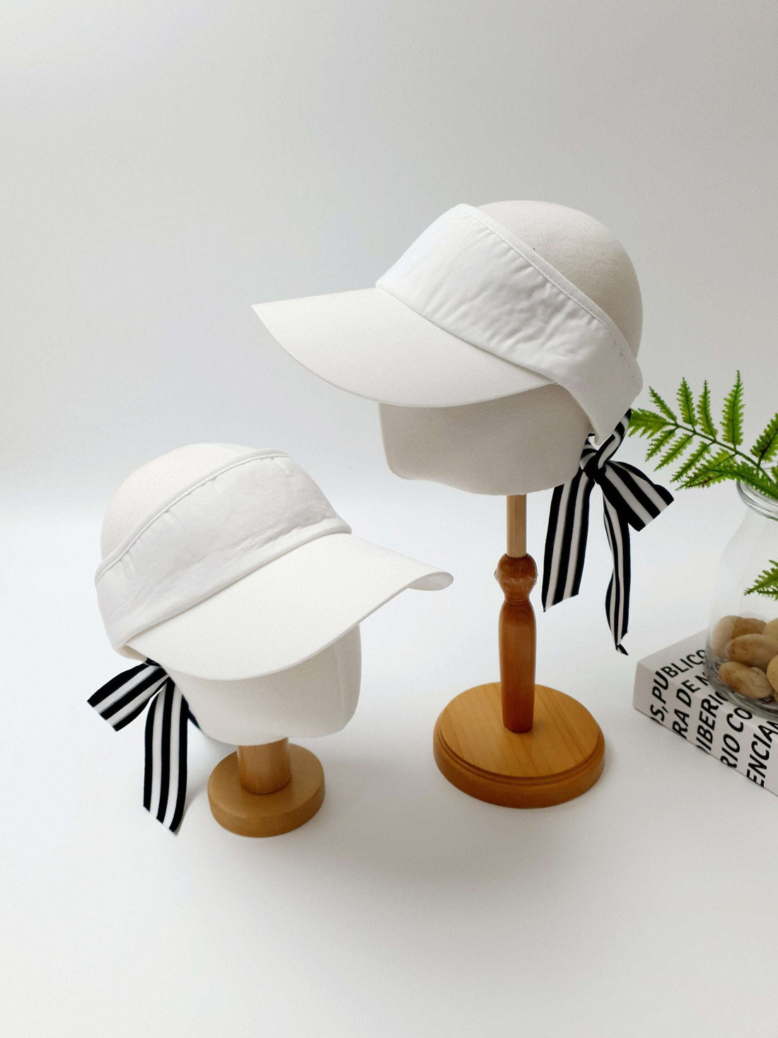 Two white lic visor hats on mannequin heads, each adorned with a stylish black and white striped ribbon tie.