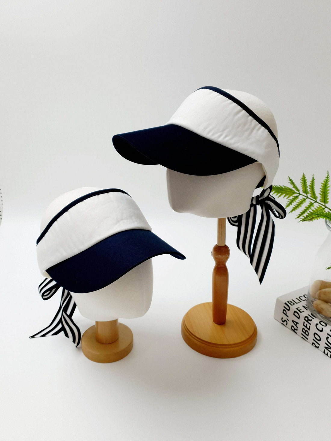 Two lic visor hats: white with navy brim and striped ribbon tie detail, displayed on mannequin heads.