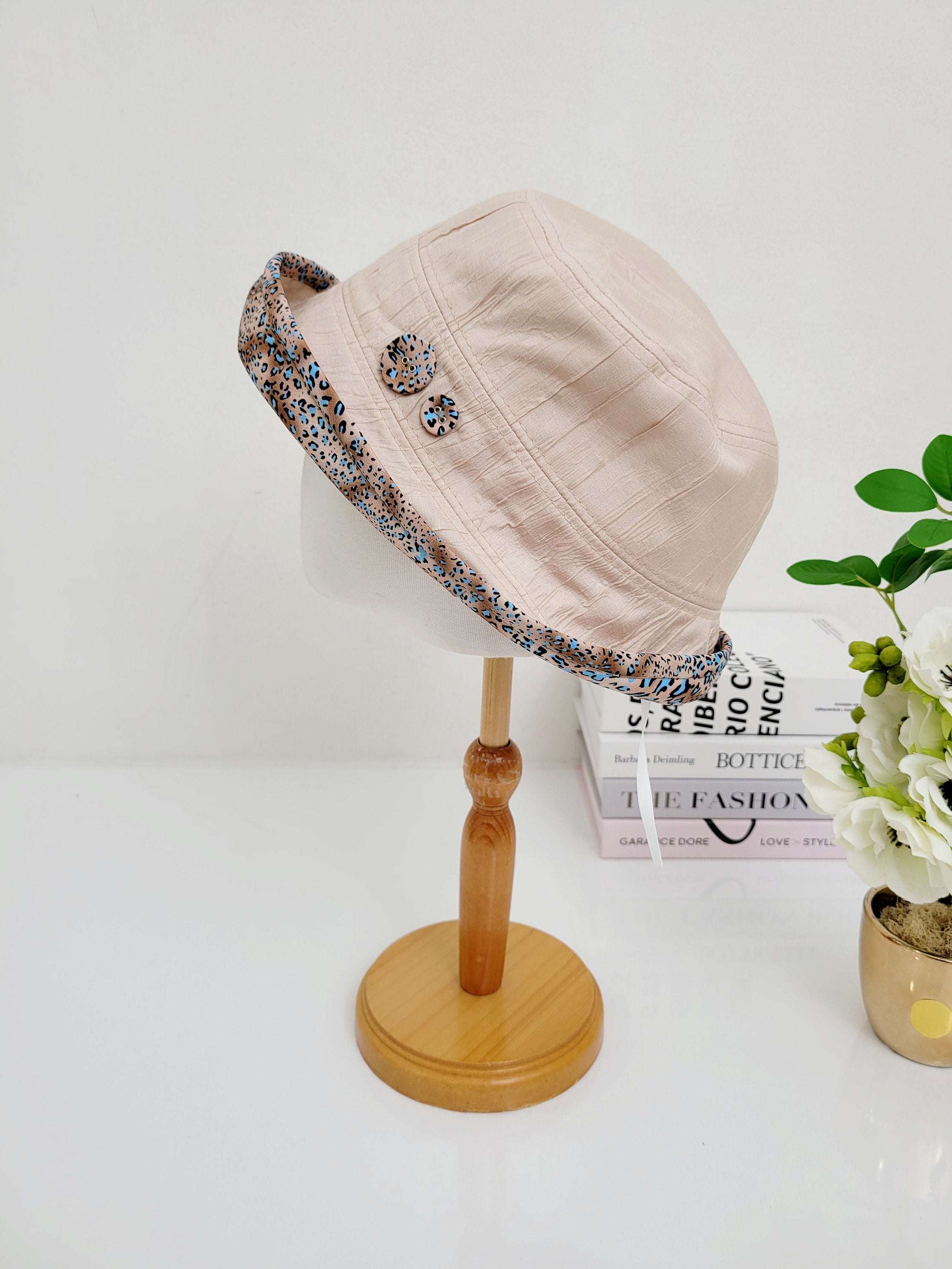 LEOPARD LINEN BUCKET-FOLDERABLE: Beige linen bucket hat with a stylish leopard print brim and decorative buttons, displayed on a wooden stand.