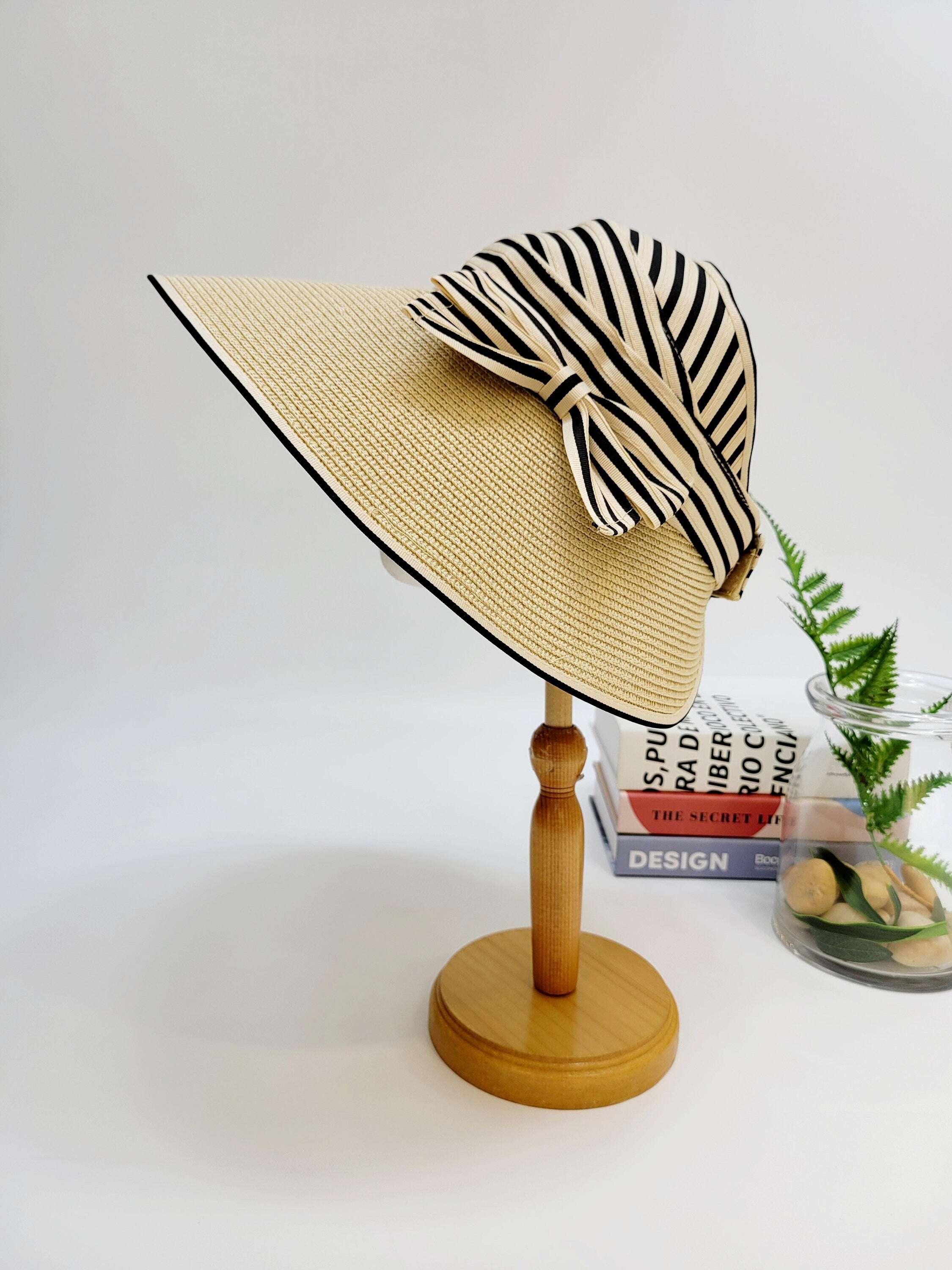 MONROE hat: A beige straw sun visor with black trim, adorned with a black and white striped bow, displayed on a wooden stand.