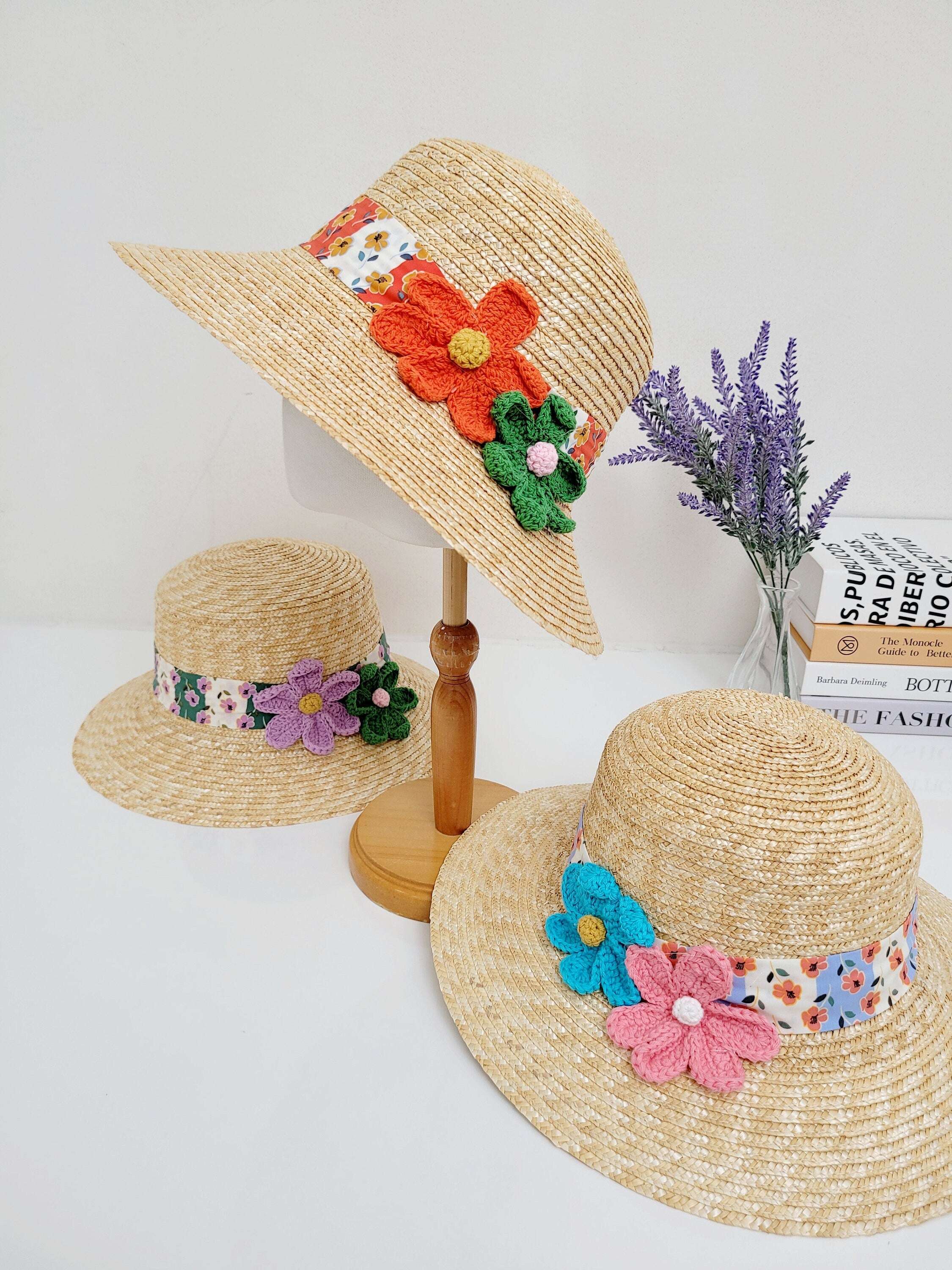 Three daisy - straw bucket hats with floral bands and crochet flowers. One hat is on a wooden stand. The straw bucket hats feature colorful floral accents.