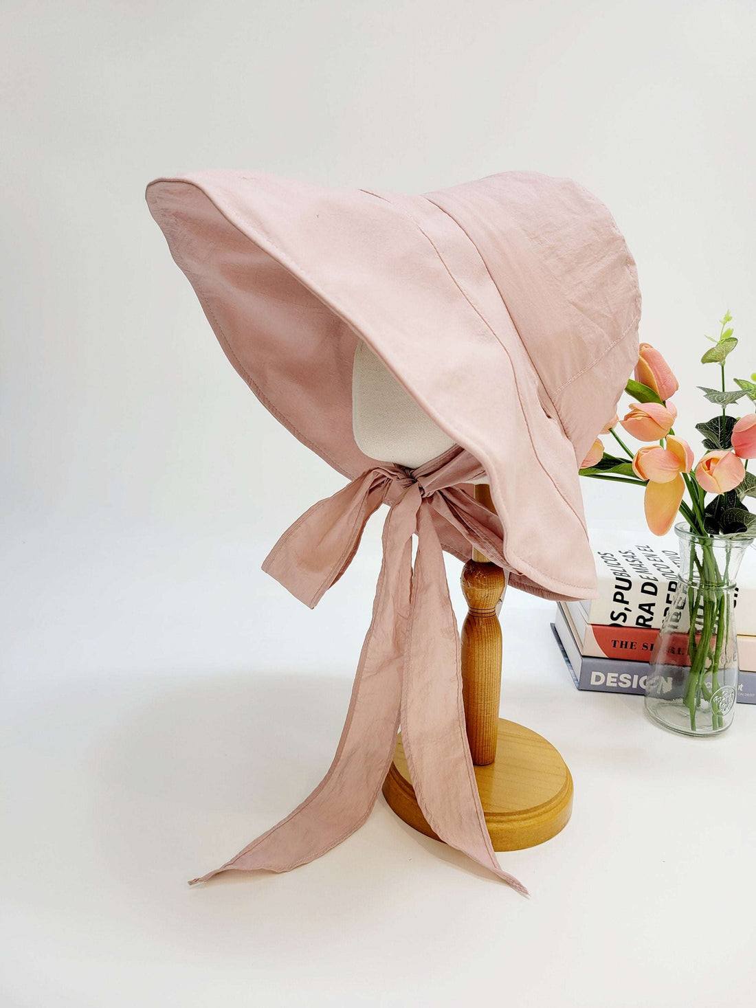 my fair lady: Soft pink sun hat with a wide, floppy brim and long ties, displayed on a stand with flowers in the background.