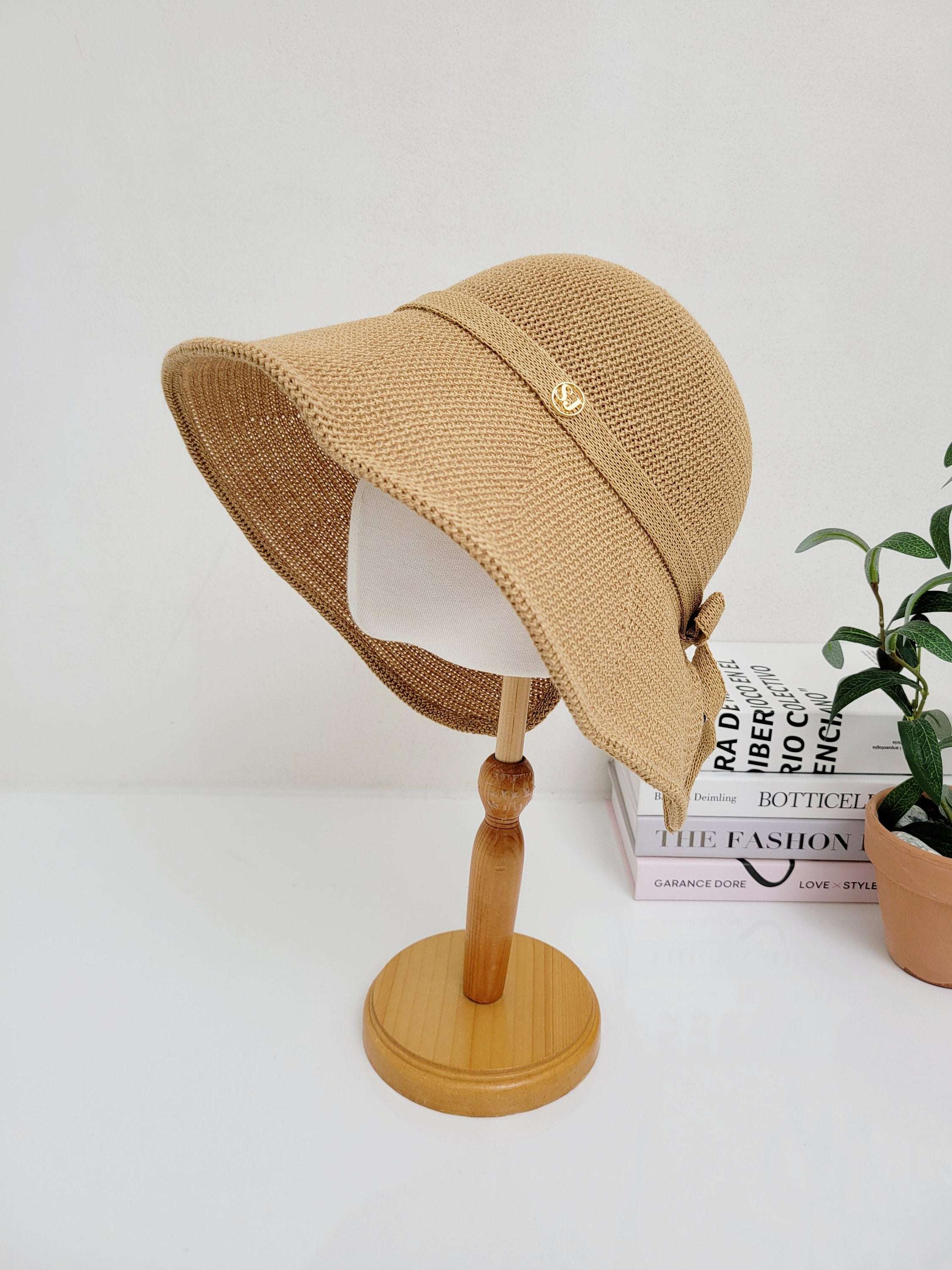 Tan woven sun hat on a wooden stand, featuring a ribbon detail and a small gold embellishment. Perfect for sun protection and stylish accessorizing.