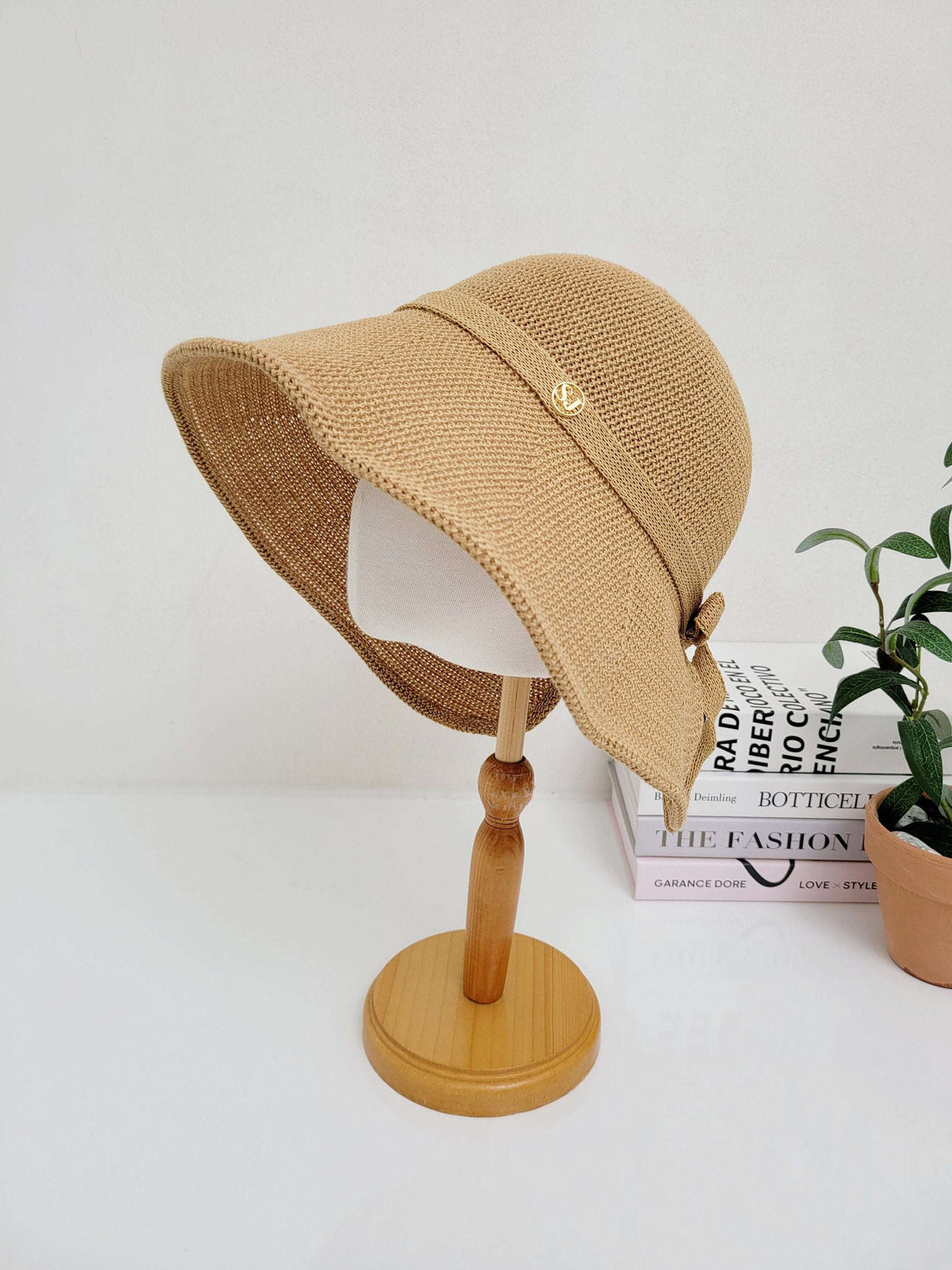 Tan woven sun hat on a wooden stand, featuring a ribbon detail and a small gold embellishment. Perfect for sun protection and stylish accessorizing.