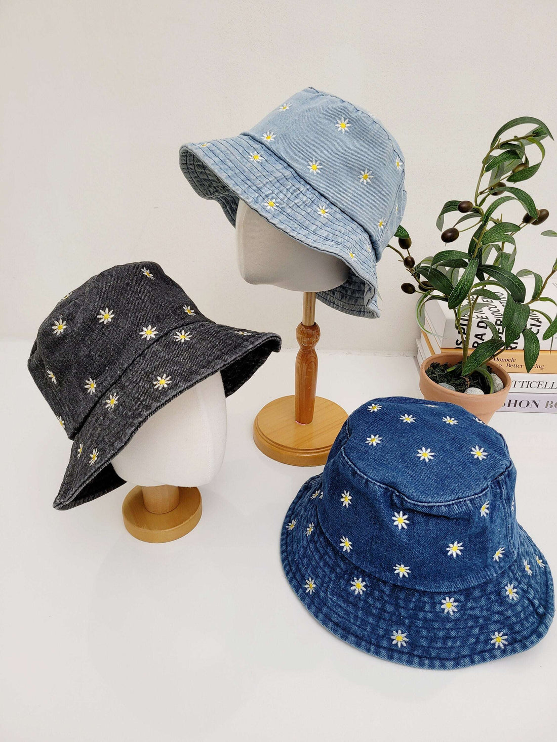 Three daisy denim bucket hats: light blue on a stand, dark blue flat, and black on a stand, all with embroidered white daisies.