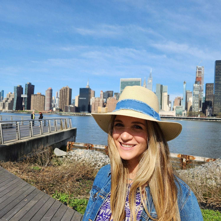 Woman wearing a long island straw hat with a blue band, denim jacket, and patterned dress, smiling with a city skyline in the background.
