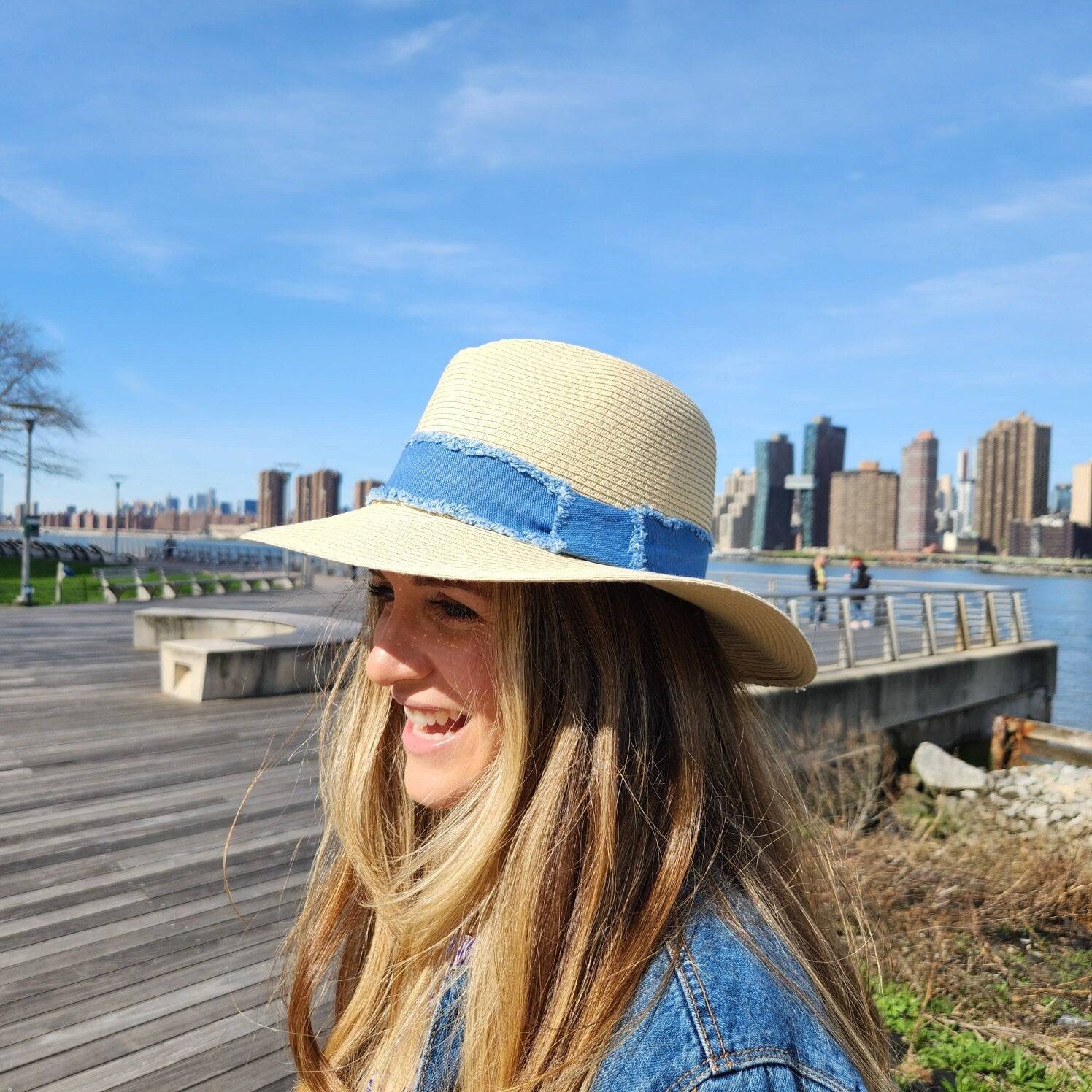 
long island straw hat with a blue denim band, worn outdoors on a sunny day with a city skyline in the background.
