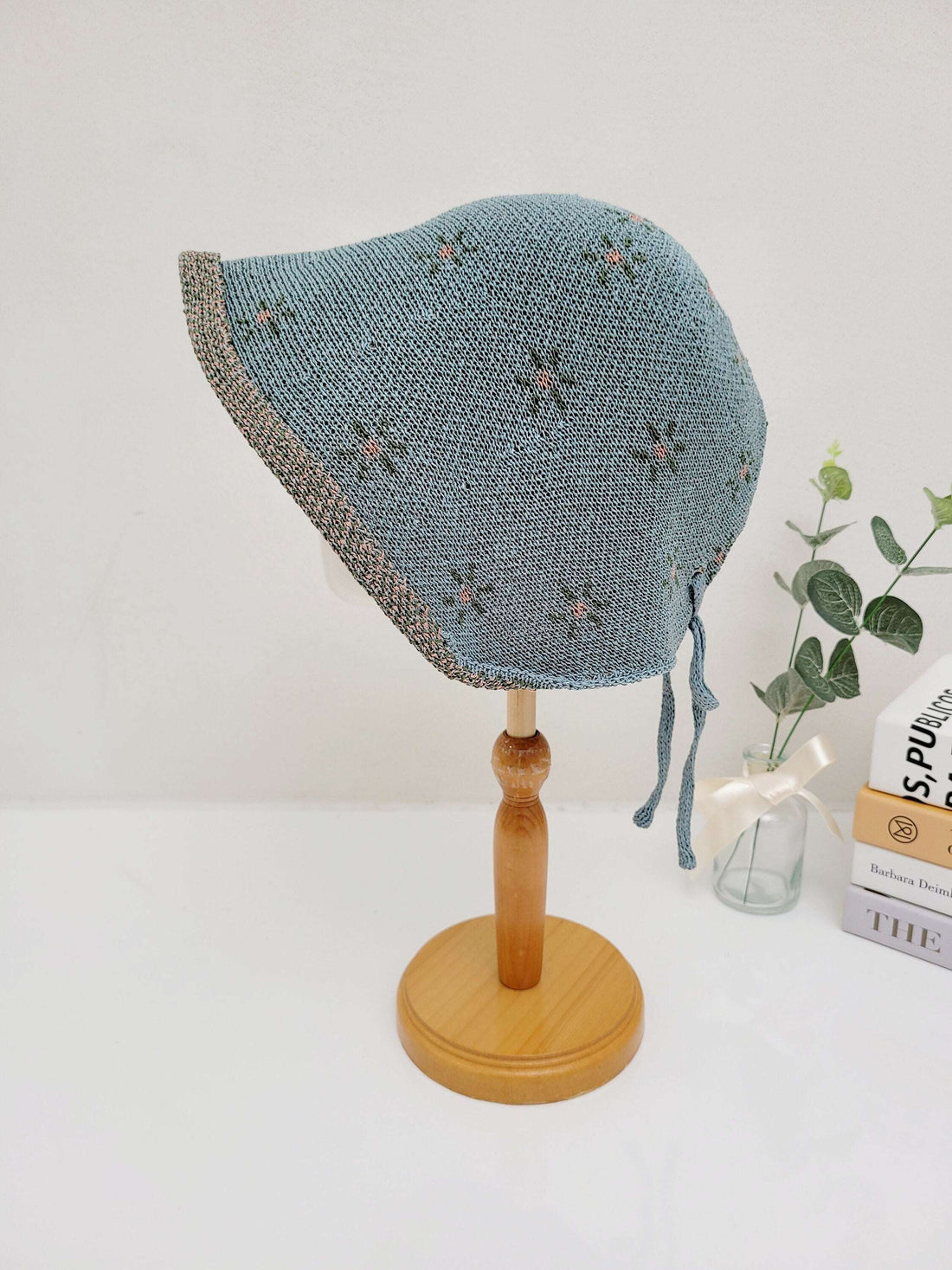 Blue knit emma bonnet with delicate floral embroidery, displayed on a wooden stand. Features tie closures and a brim.