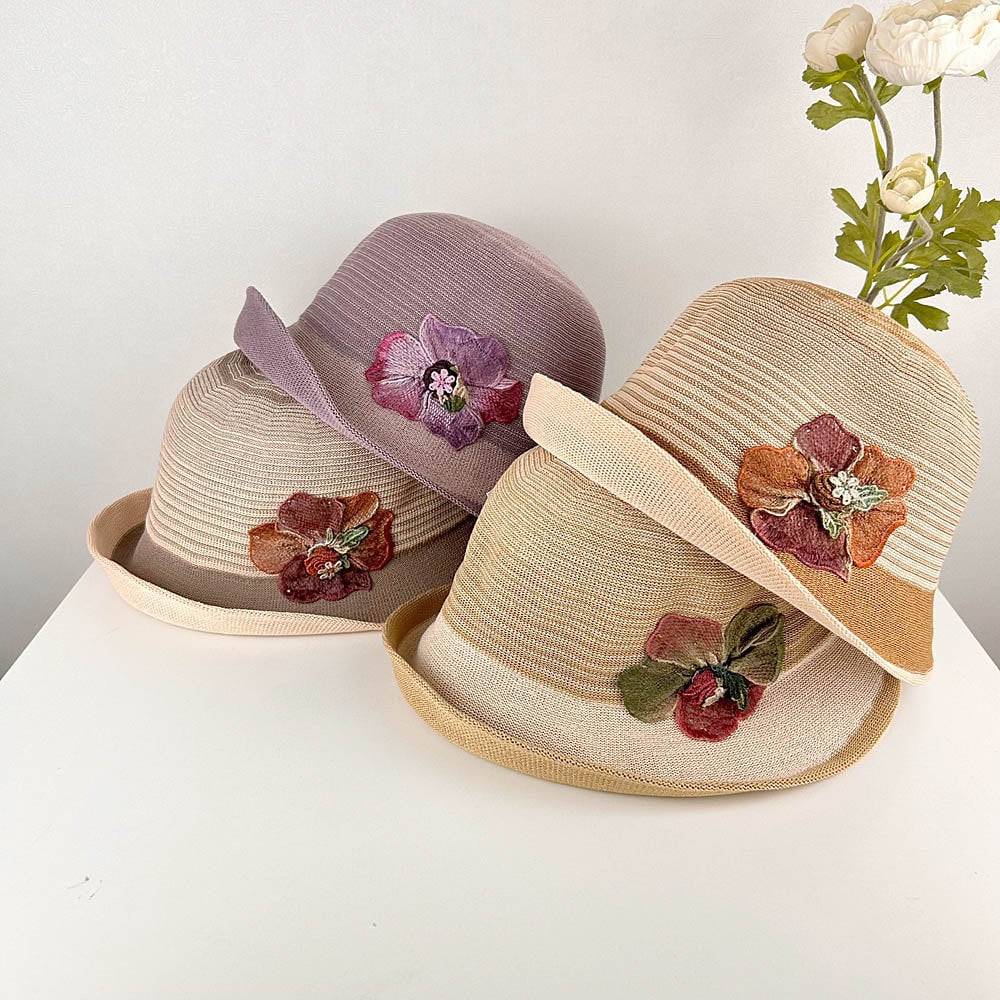 Three straw-like floral crown sun hats in beige, tan, and lavender, each adorned with unique fabric flower embellishments. 