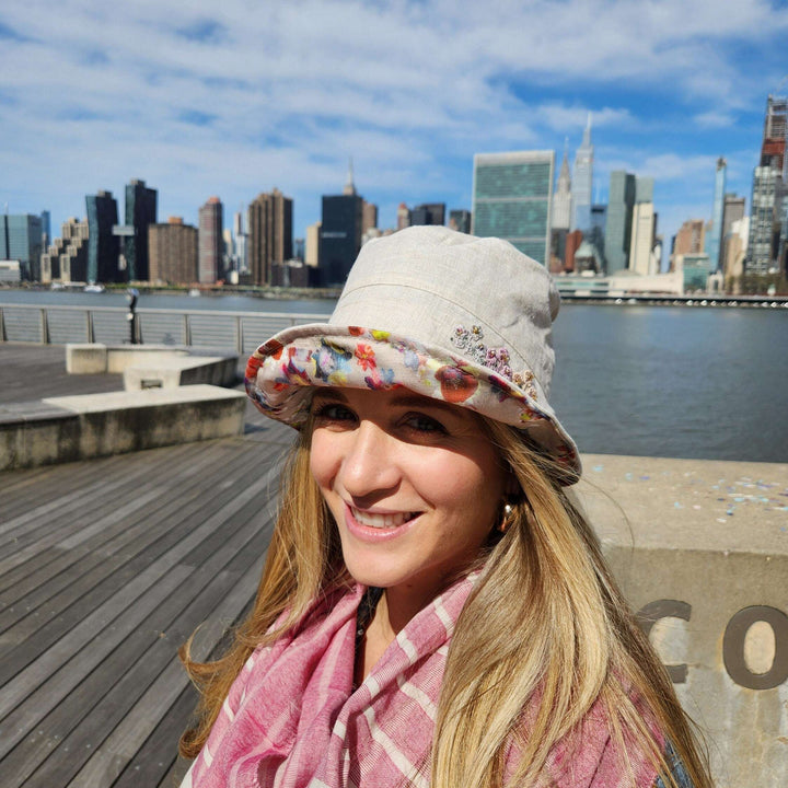 Woman smiles wearing a floral linen bucket-folderable hat with a pink scarf; cityscape backdrop.