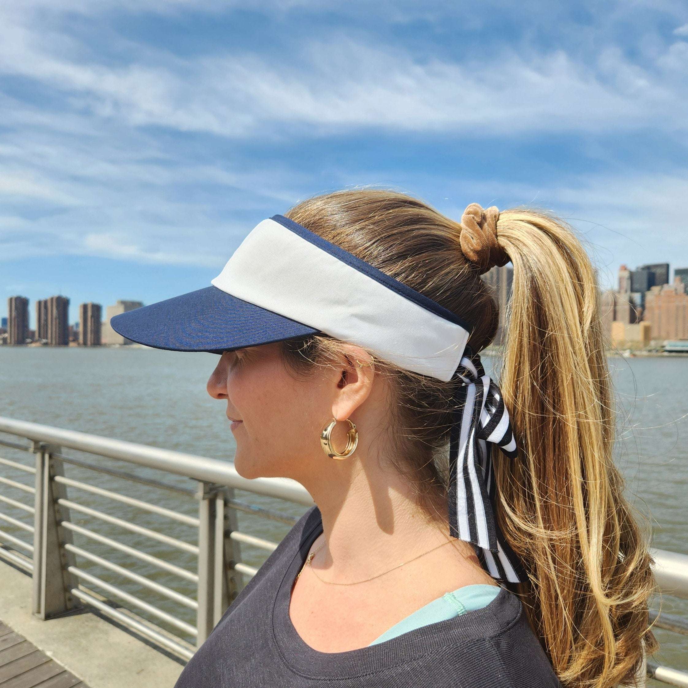 Navy and white lic visor with a striped ribbon detail, worn with a blonde ponytail. Cityscape backdrop.