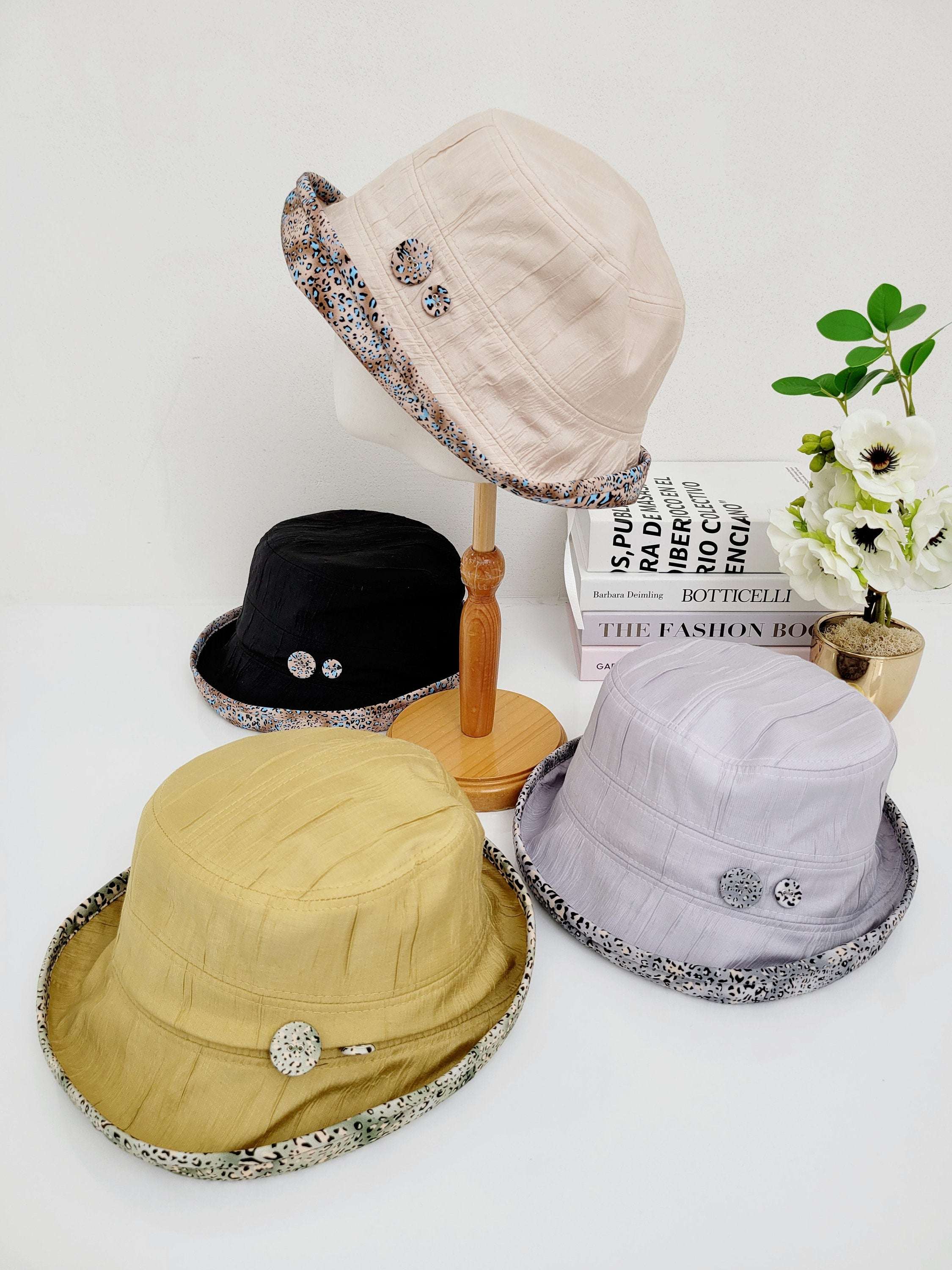 Four leopard linen bucket-folderable hats: beige, black, gold, and lavender, each with a patterned brim and decorative button detail.