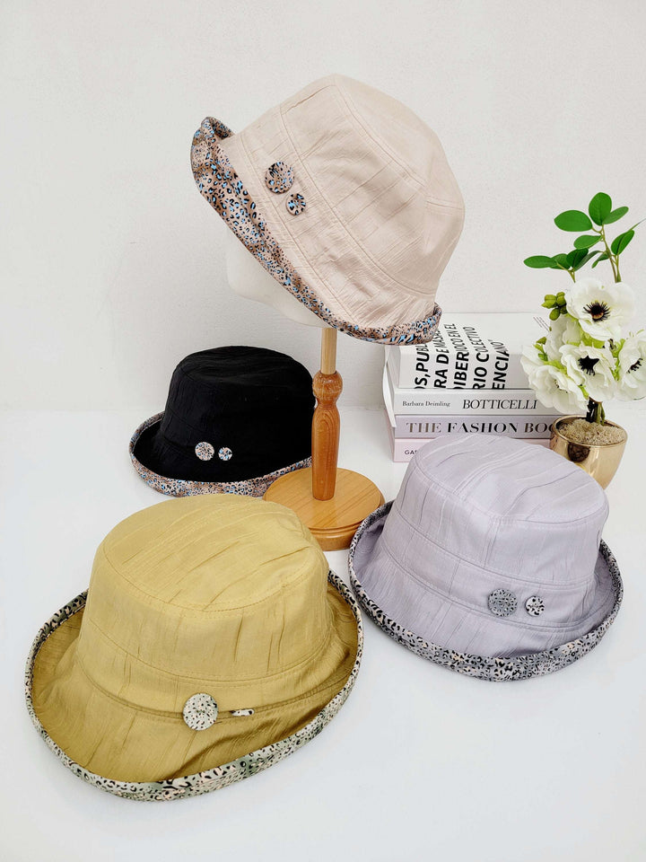 Four leopard linen bucket-folderable hats: beige, black, gold, and lavender, each with a patterned brim and decorative button detail.