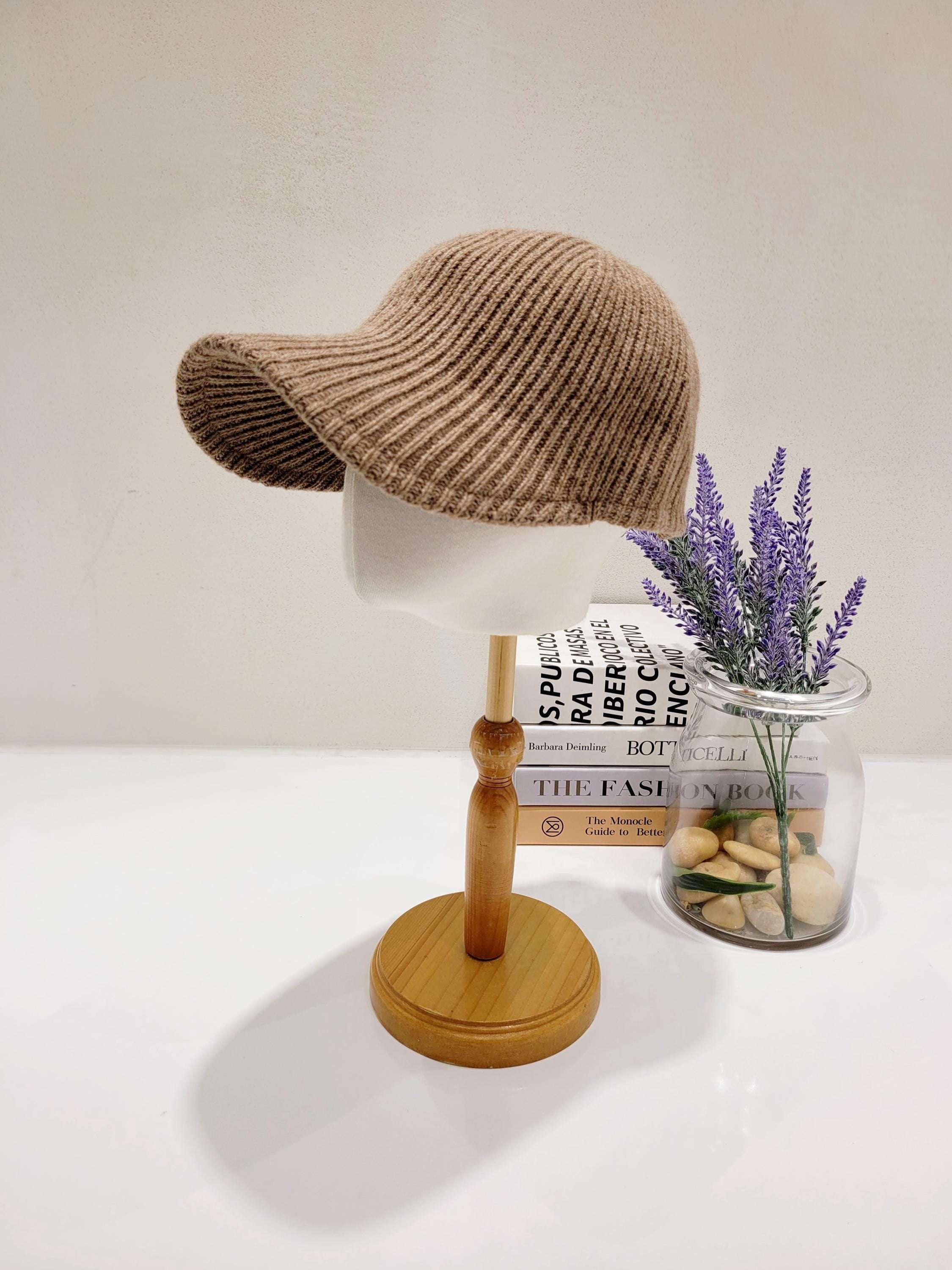 Brown wool ribbed cap displayed on a wooden stand, showcasing its textured knit and stylish brim. Beside it, books and lavender add a touch of elegance.