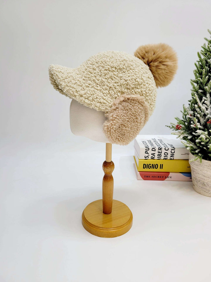 WOOF WOOF: Beige sherpa hat with fuzzy ear flaps and a fluffy brown pompom, displayed on a wooden stand.