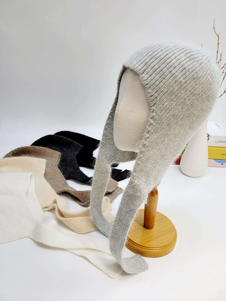 Gray knit wool balaclava on a mannequin, featuring long ties. Other colors (white, beige, brown, black) are visible in the background.