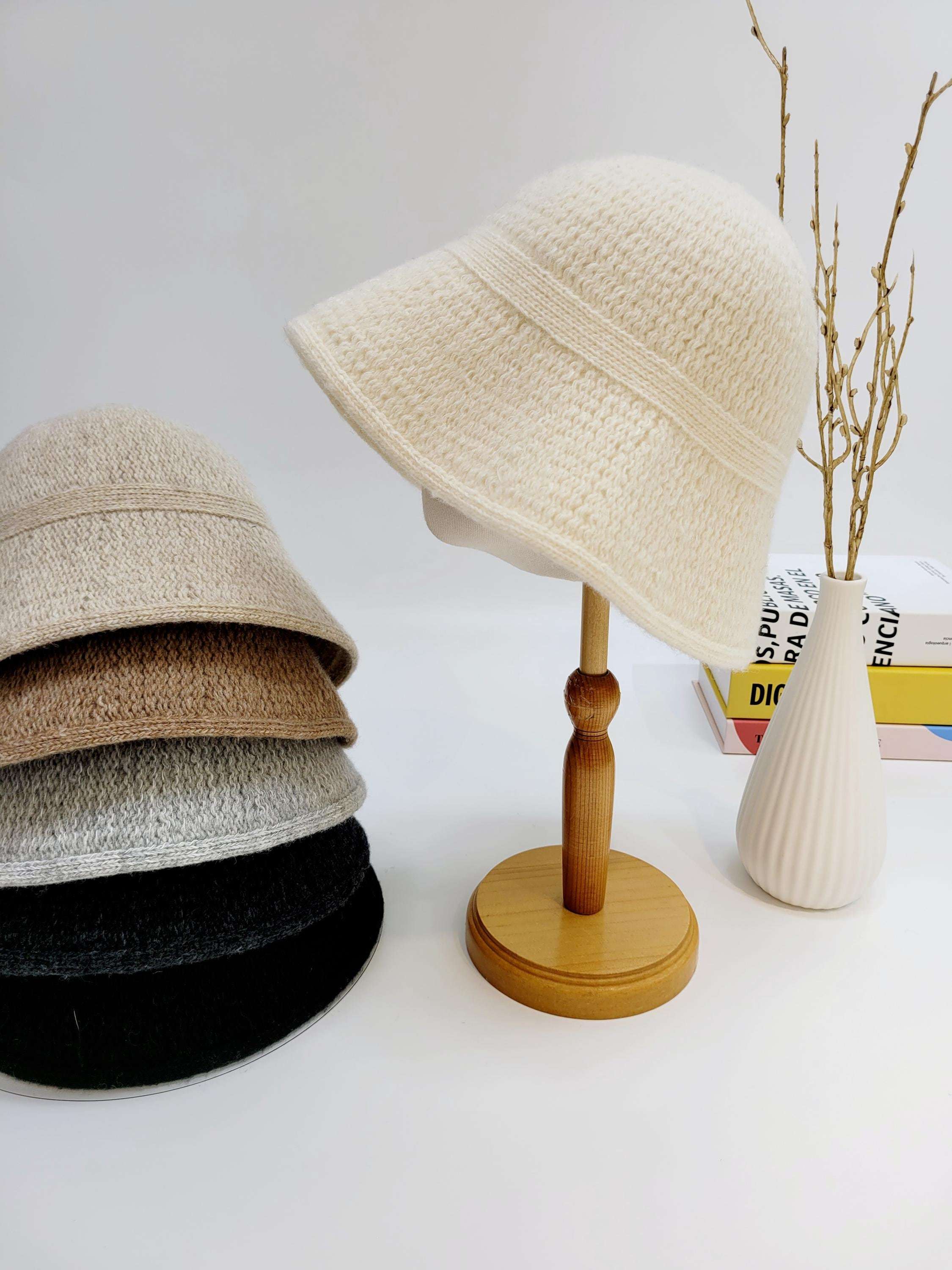 Stack of knitted victoria's winter hat in beige, brown, grey, and black, with a single cream hat on a wooden stand next to a vase.