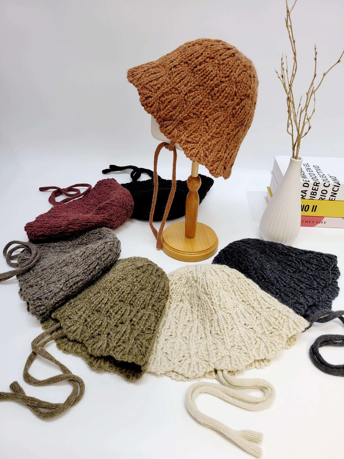 Assorted braided wool hat collection in brown, burgundy, gray, olive, cream, and charcoal; one on a wooden display stand.