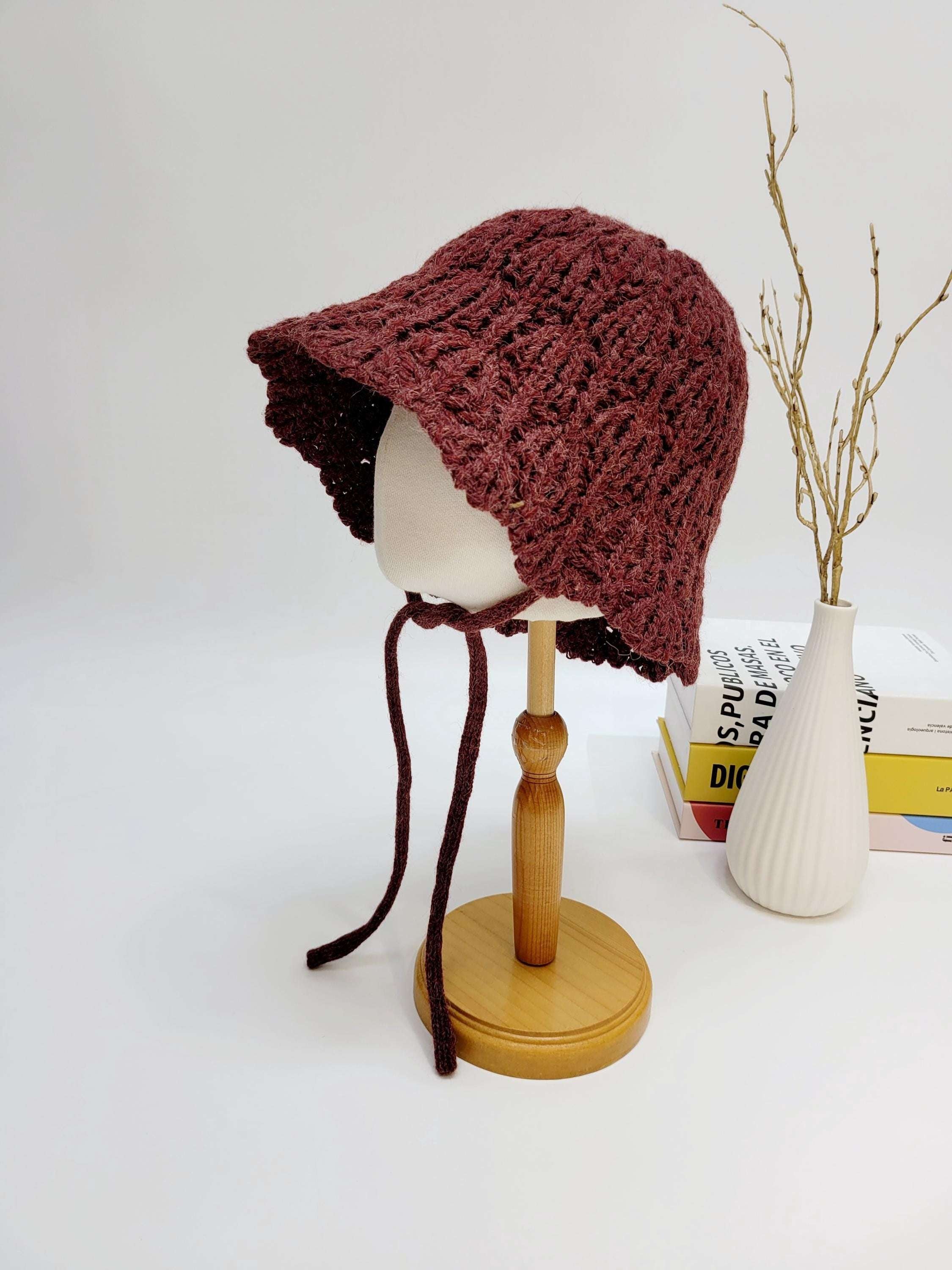 Burgundy braided wool hat baby bonnet with tie strings, displayed on a wooden stand