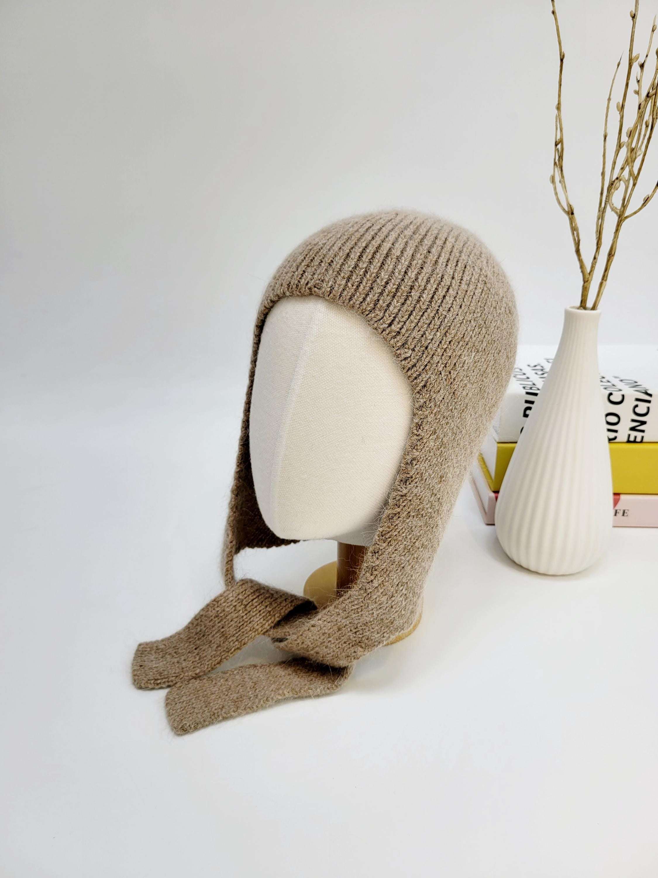 Beige wool balaclava on a mannequin head, featuring a ribbed knit and ear flaps with tie closures.